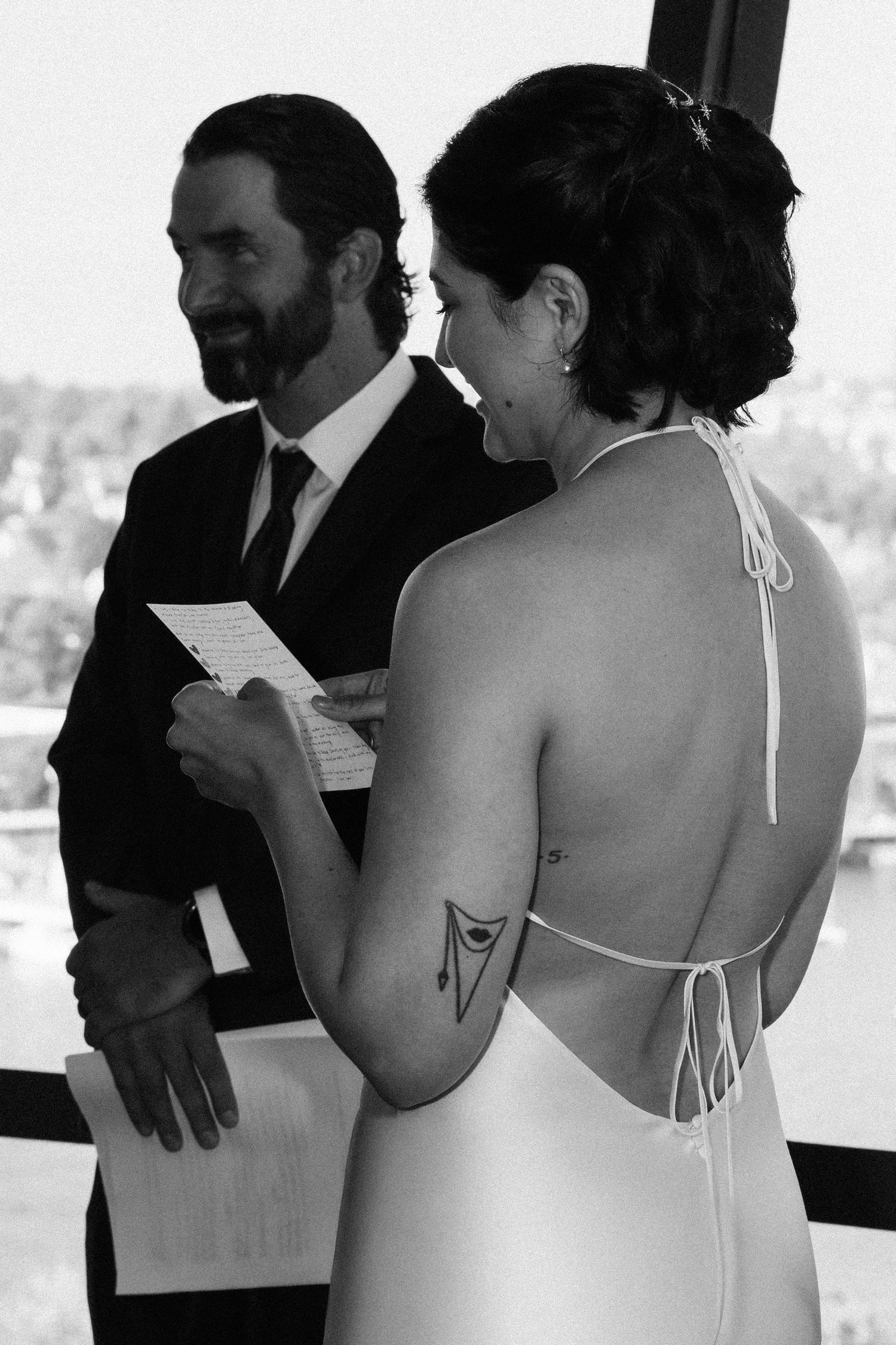 Black and white wedding photo of bride reading vows in backless halter gown with tattoo, groom in suit smiling.
