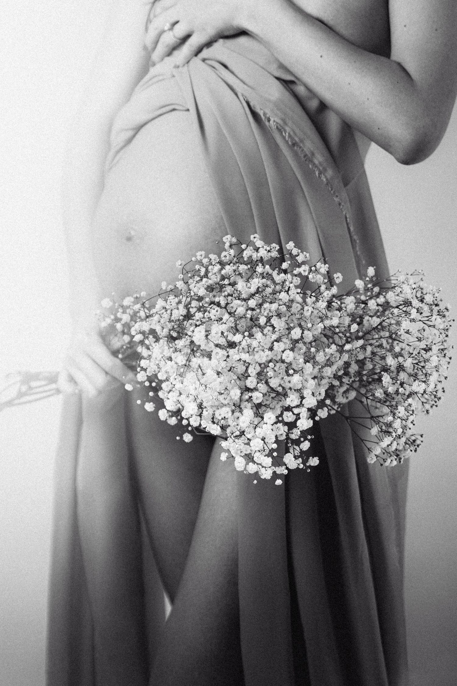 Black and white maternity photo of pregnant woman holding baby's breath flowers while draped in sheer fabric.