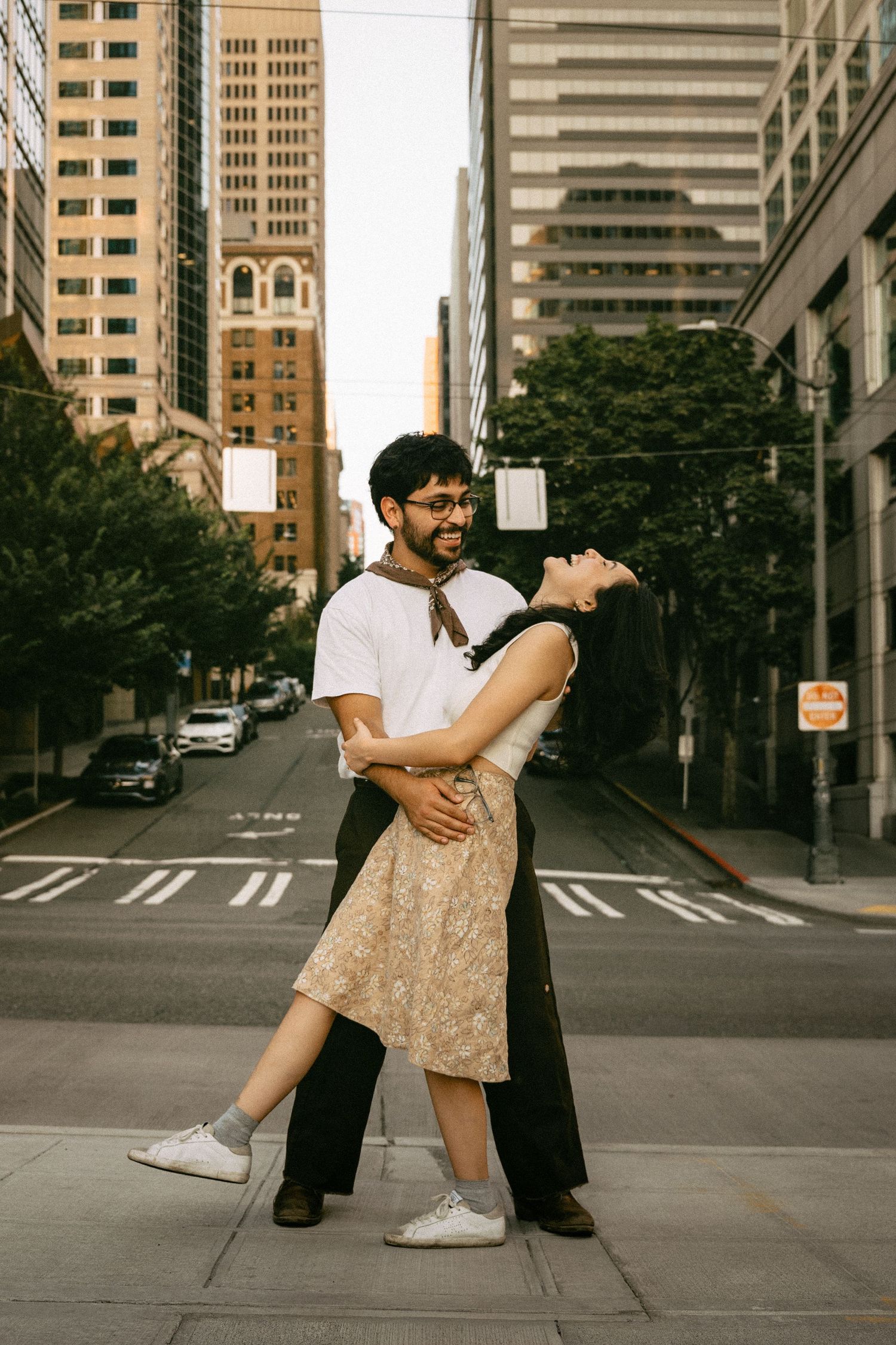 Couple dancing and embracing on a city sidewalk surrounded by tall buildings and trees on a sunny day.
