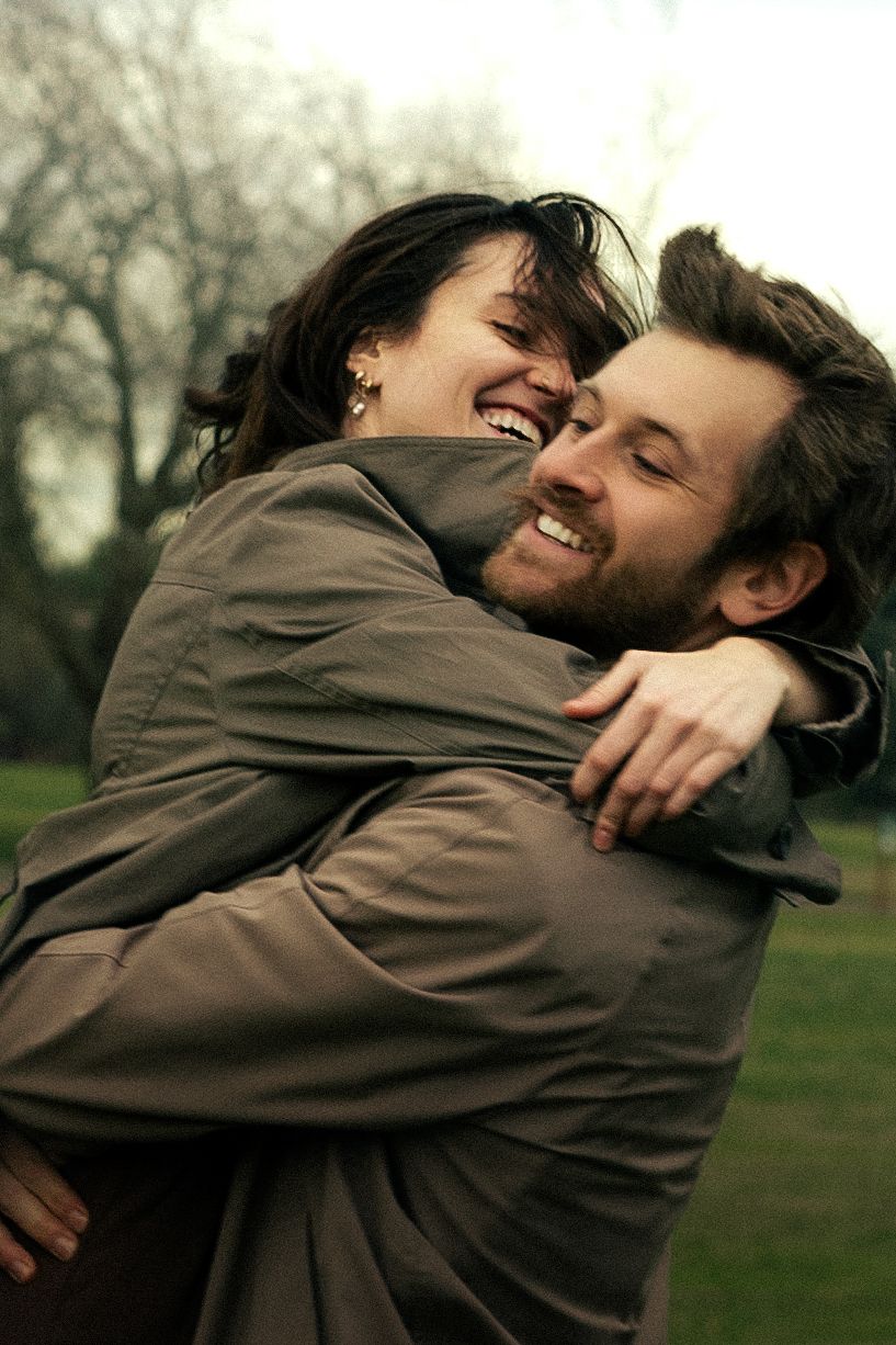 A joyful couple embraces and laughs together outdoors in a park on a cloudy day, both wearing brown jackets.