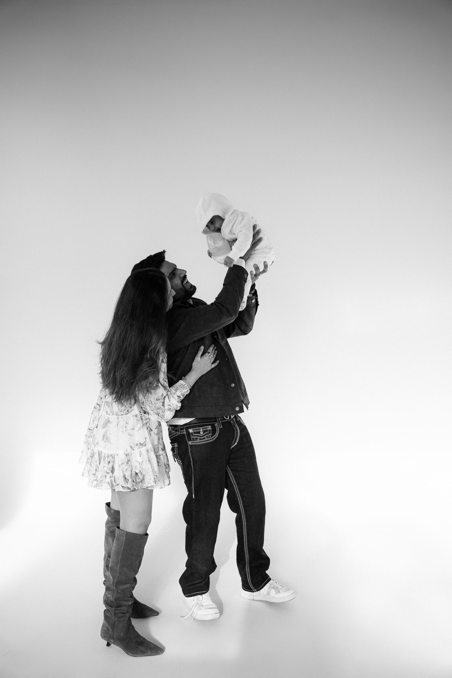 Black and white photo of a couple lifting their baby up in a bright studio setting, wearing casual stylish outfits.