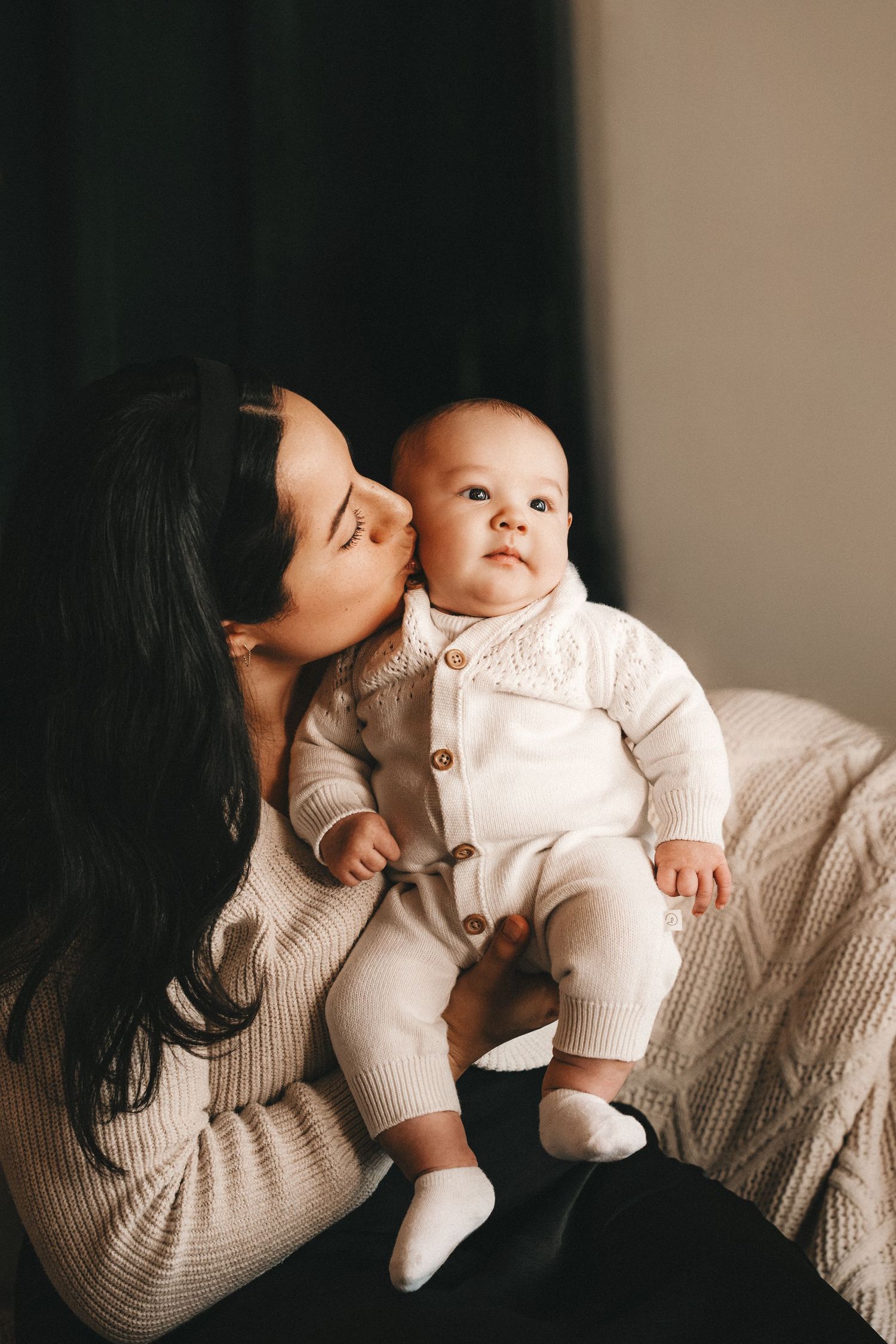 A loving mother with dark hair kisses her infant baby dressed in a cream knit outfit while sitting in a cozy chair.