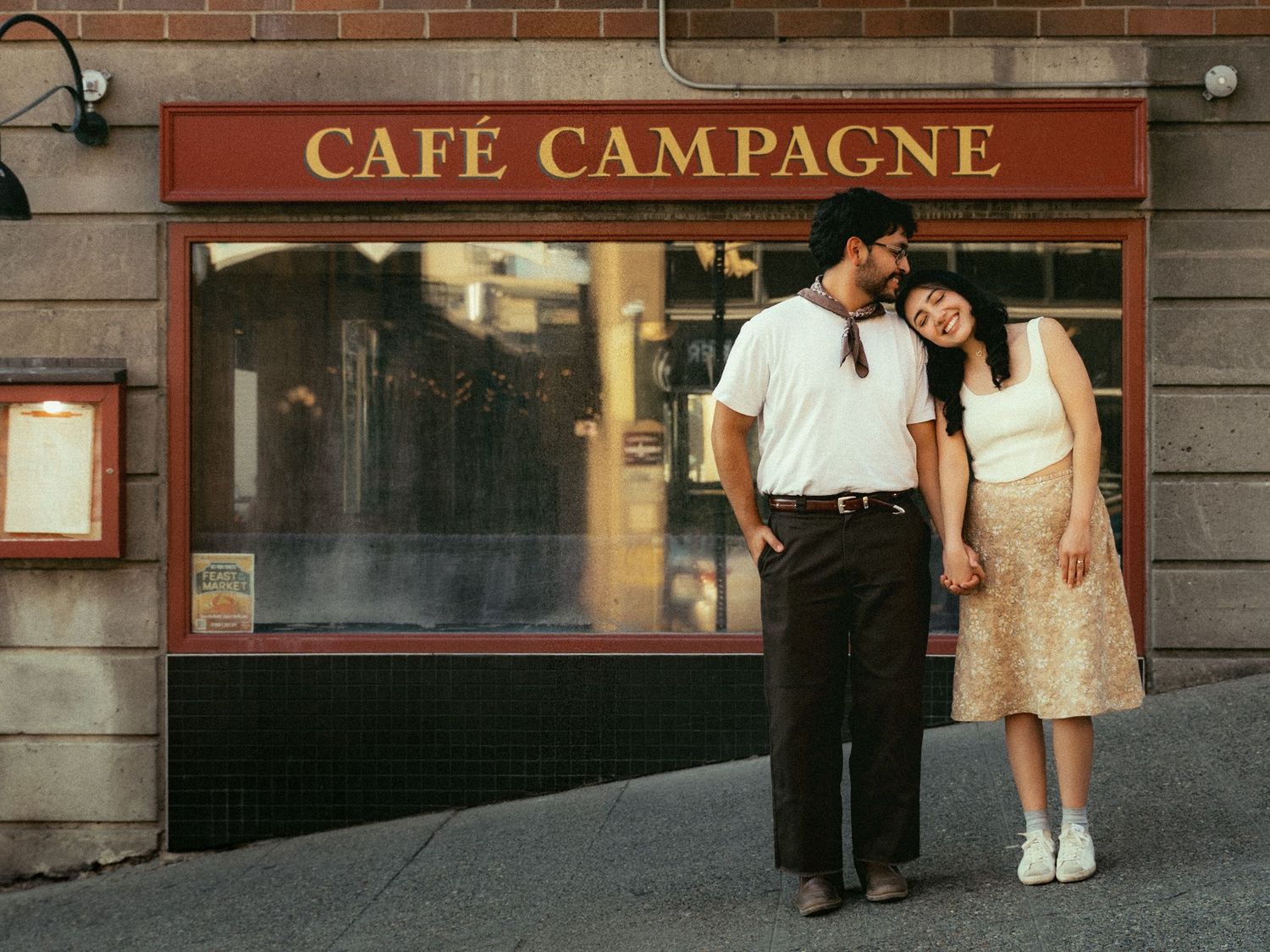 Couple holding hands and smiling outside Café Campagne restaurant on a sunny day.