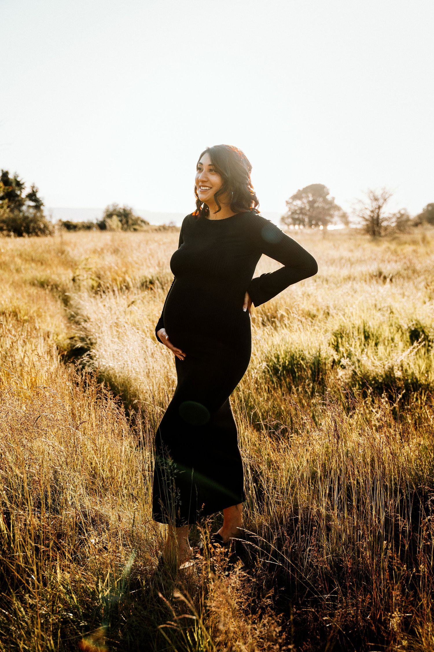 Pregnant woman in black dress standing in golden field, smiling, backlit by warm sunlight.
