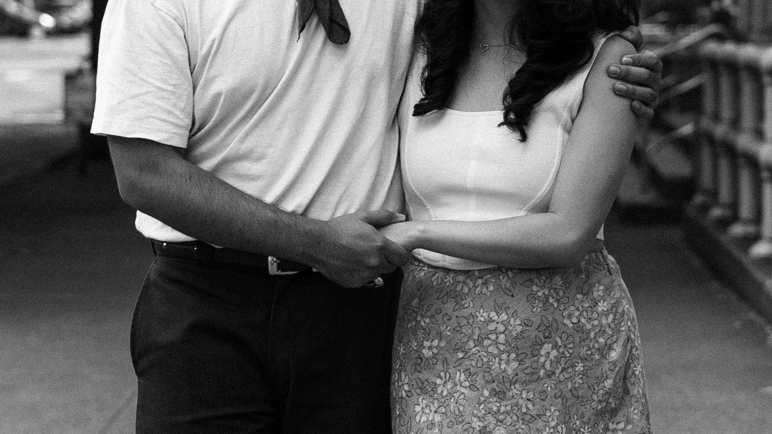 Black and white photo of a couple posing together outdoors, man in white shirt with arm around woman in floral skirt.