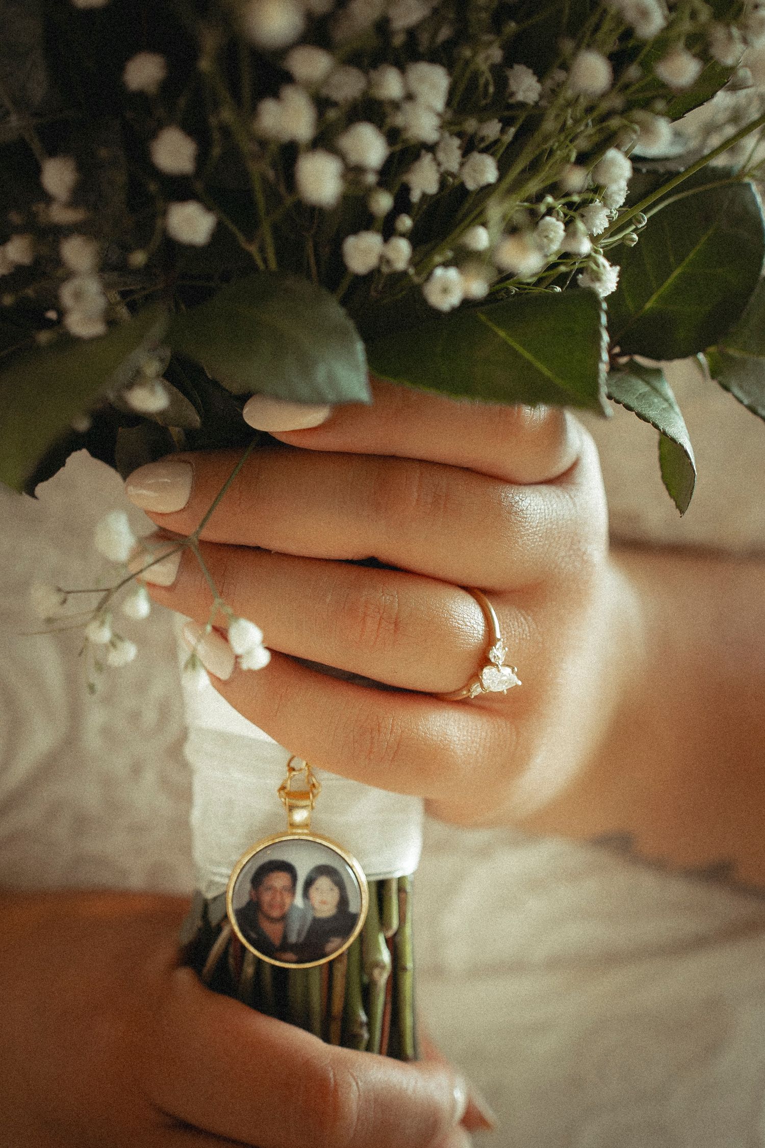 Bride holding white floral bouquet with a memorial photo charm and engagement ring on her finger.