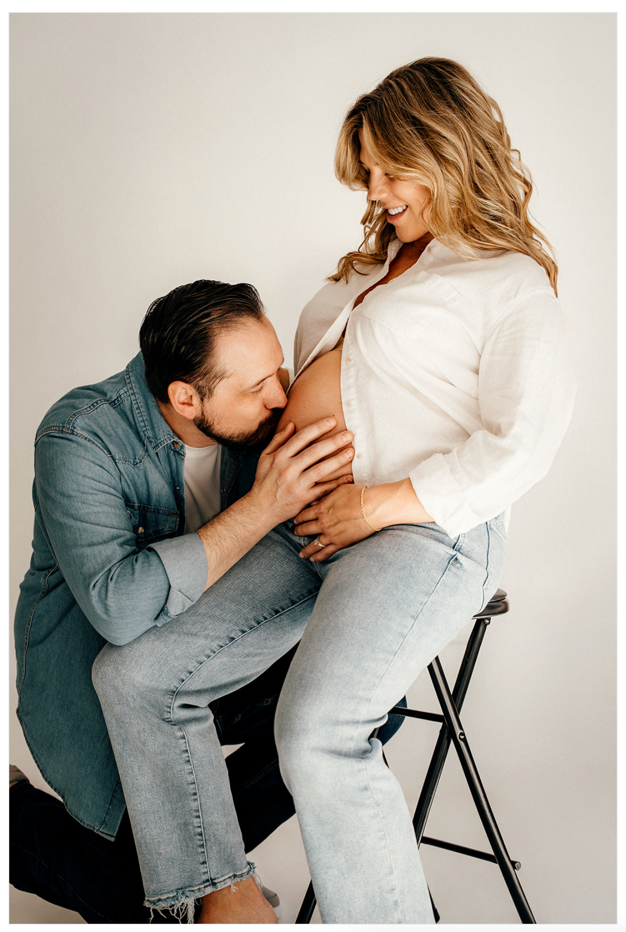 A man kisses a pregnant woman's belly while she smiles, both dressed casually in a bright studio setting.
