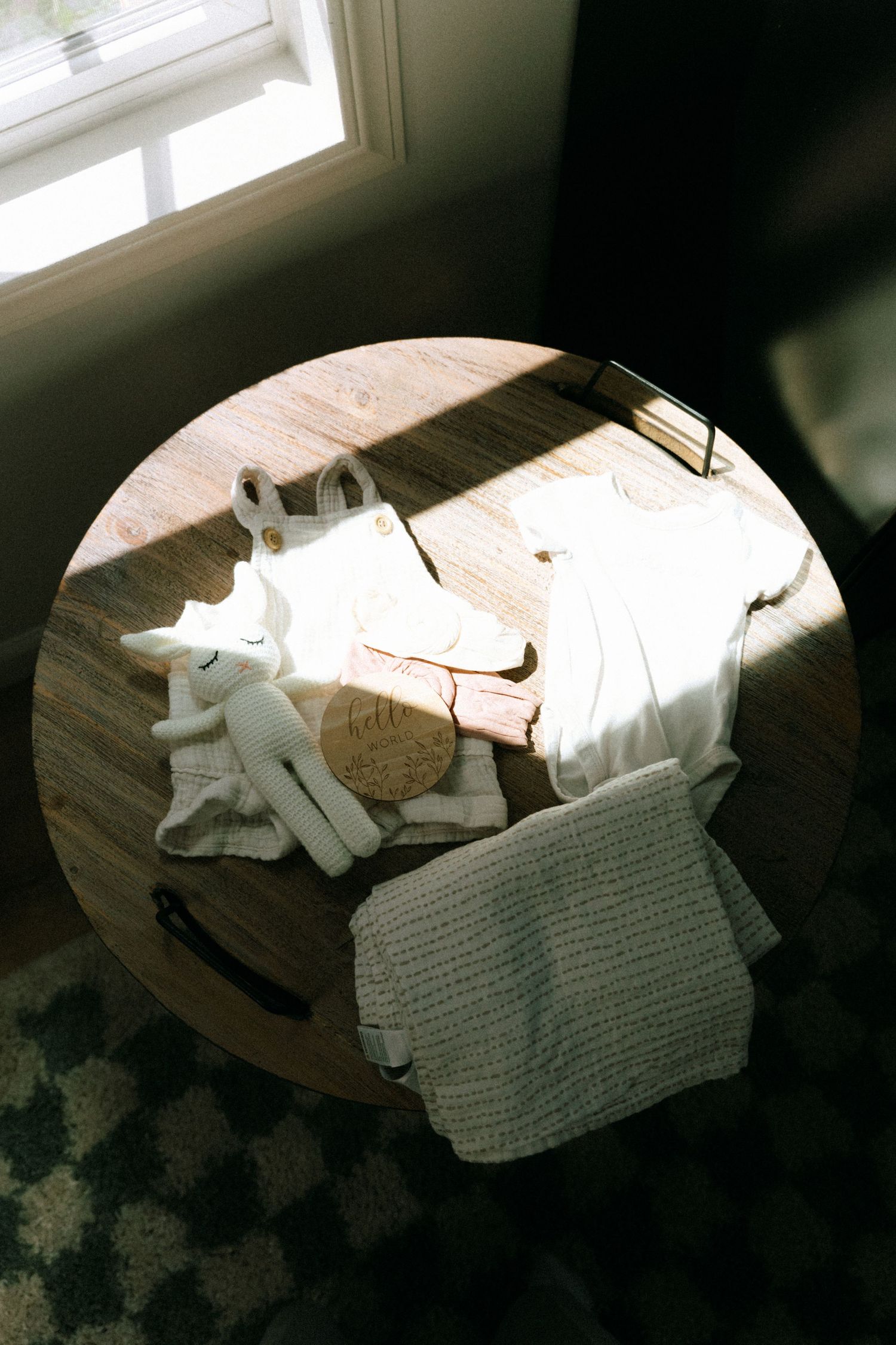 Soft white baby clothes, knit blanket, and stuffed animal toy arranged on a round wooden table in natural sunlight.