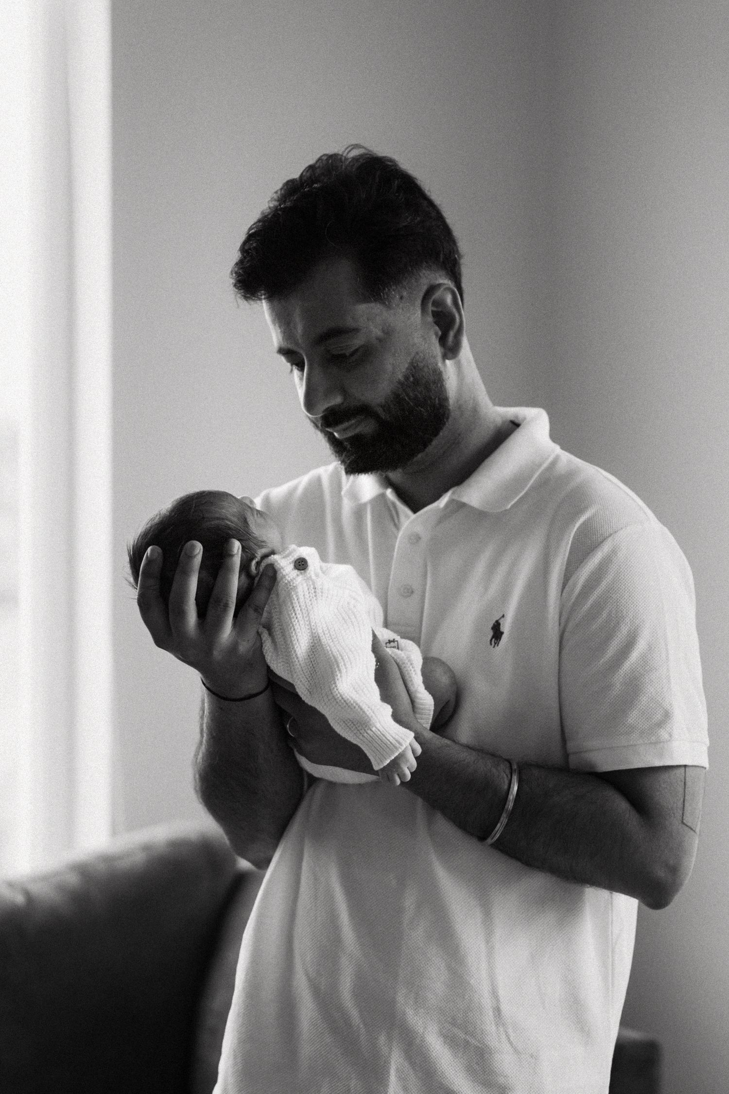 A bearded man in a white Ralph Lauren polo shirt tenderly holds a newborn baby in his arms indoors.