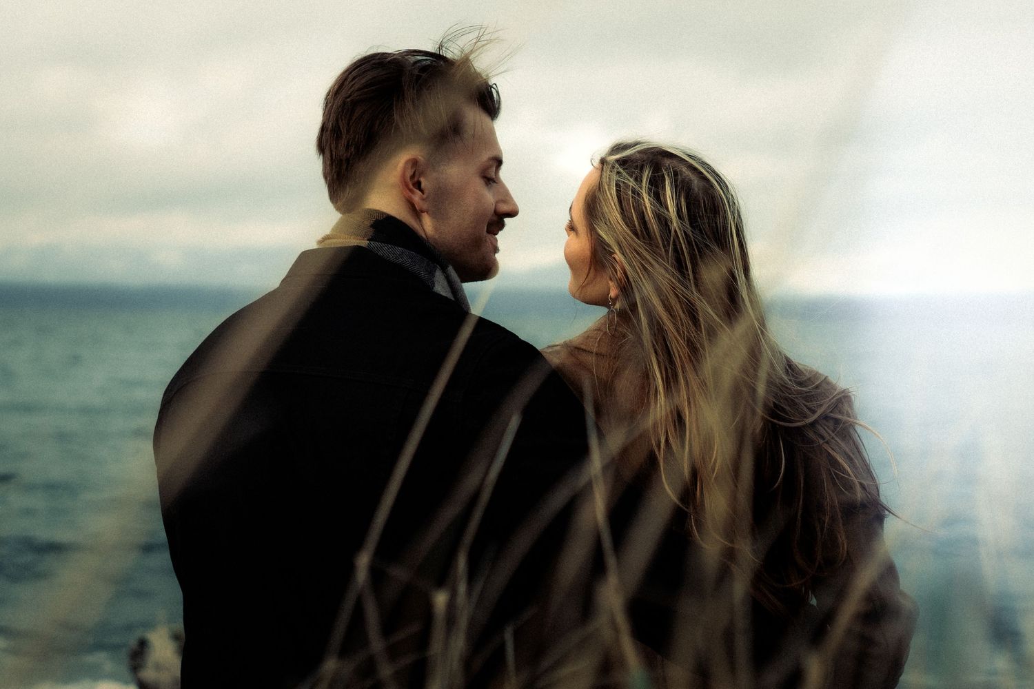 Romantic couple standing by the ocean shore, facing each other in a tender moment with dramatic cloudy sky backdrop.