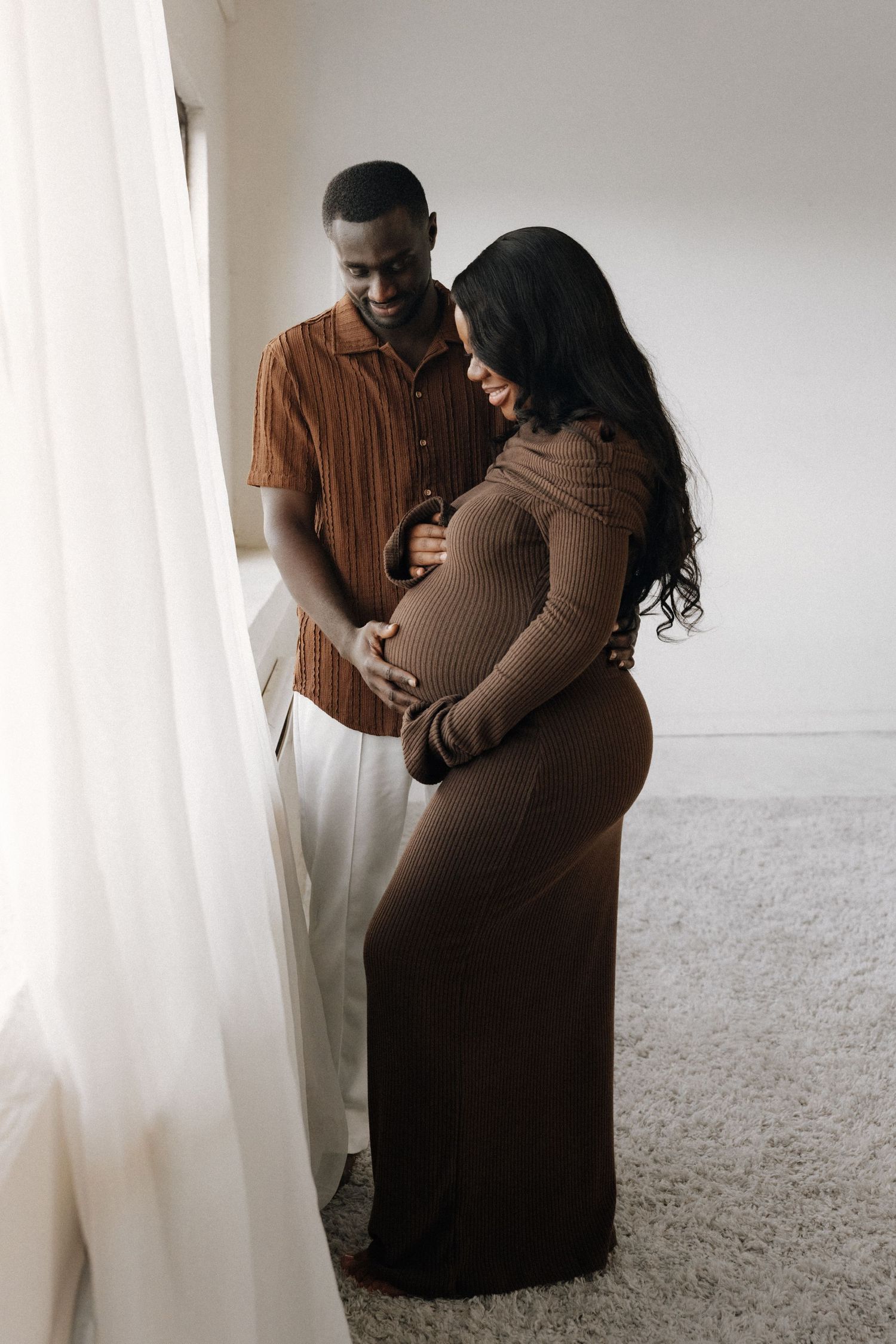 A smiling couple in matching brown outfits share a tender moment, cradling a baby bump near a sunlit window.