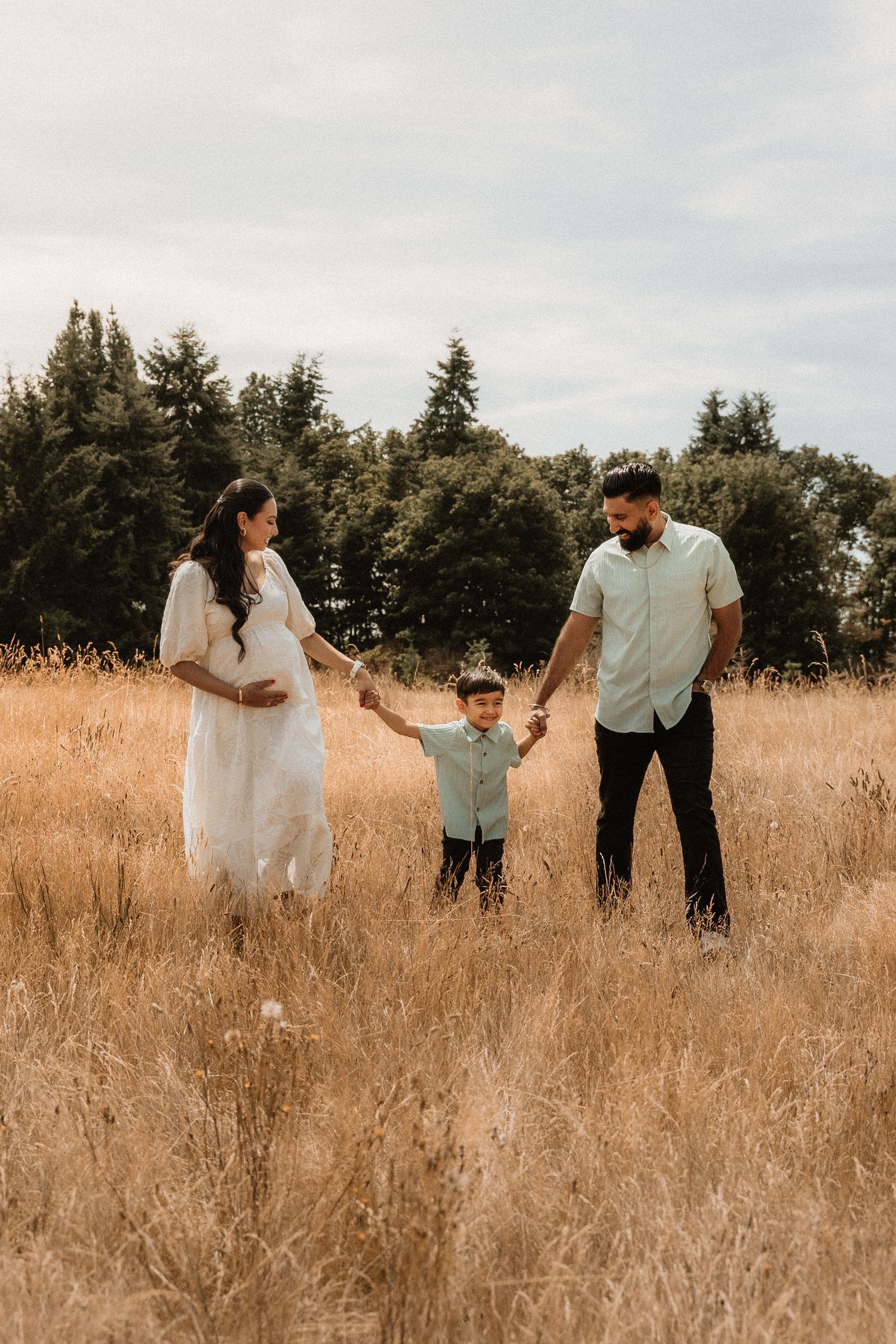 Pregnant woman in white dress and man hold hands with young boy in golden field with evergreen trees.