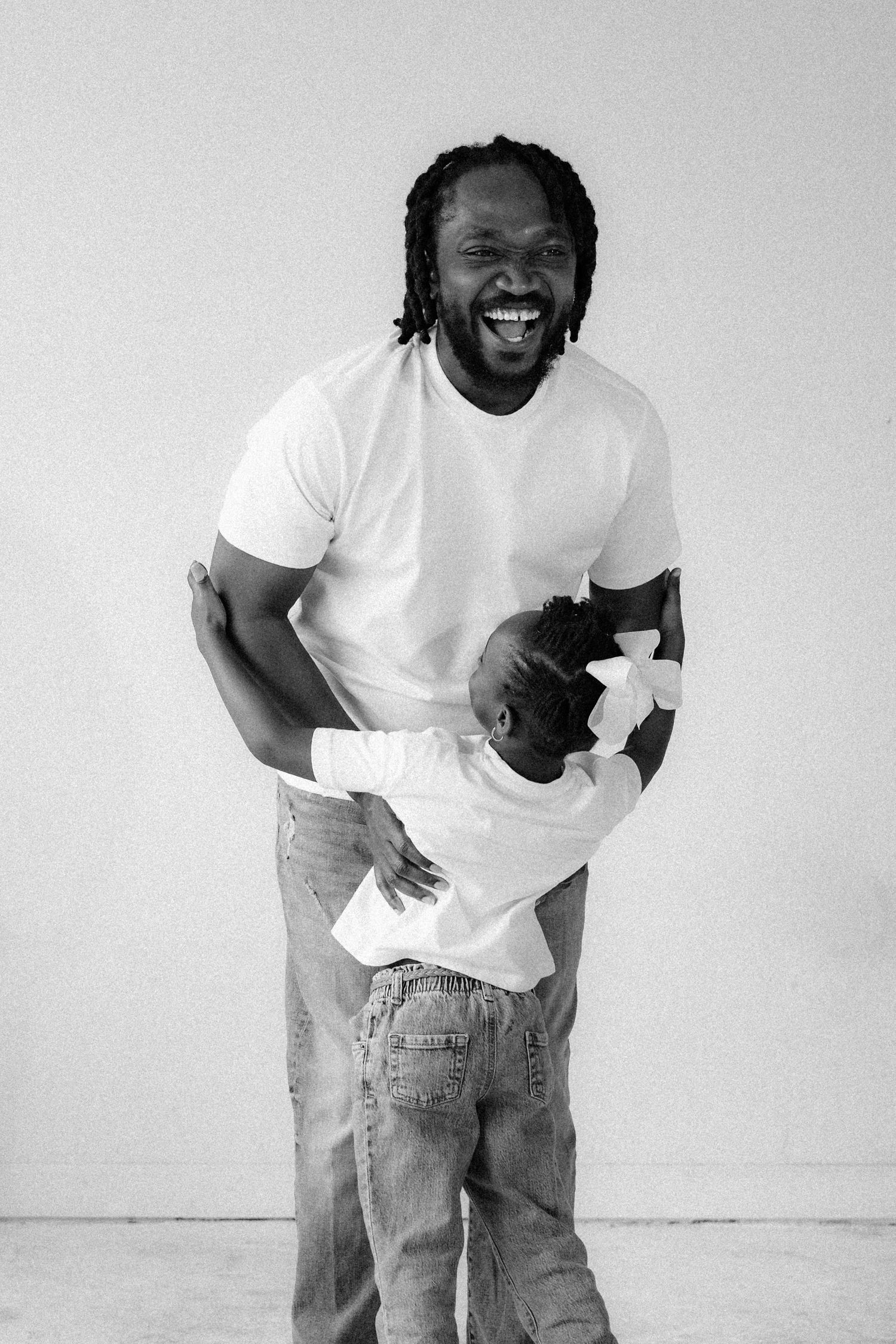 Black and white photo of a laughing man with dreadlocks holding a baby in a white shirt and jeans.