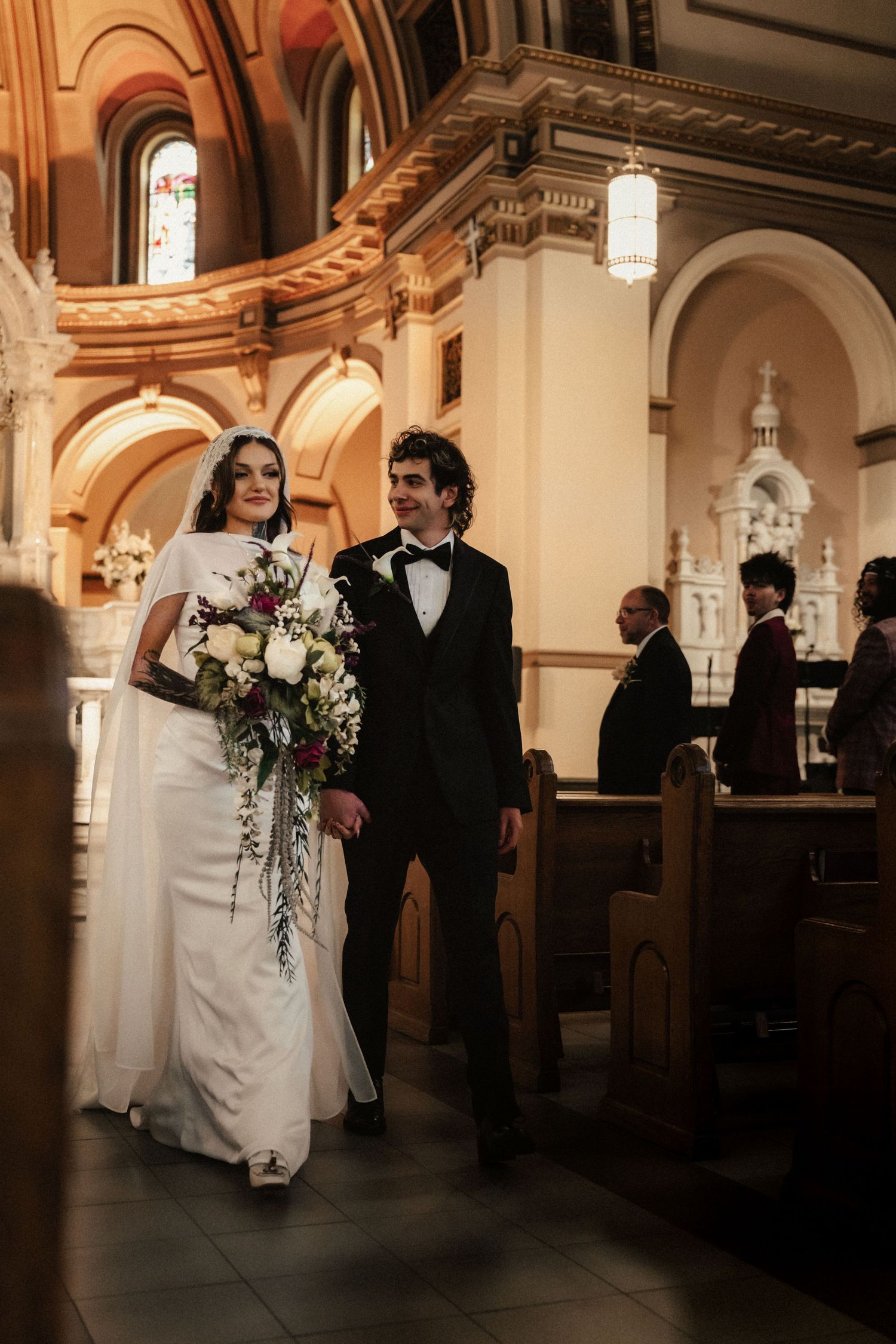 Bride and groom walk down church aisle after wedding ceremony, bride holding cascading white and purple bouquet.