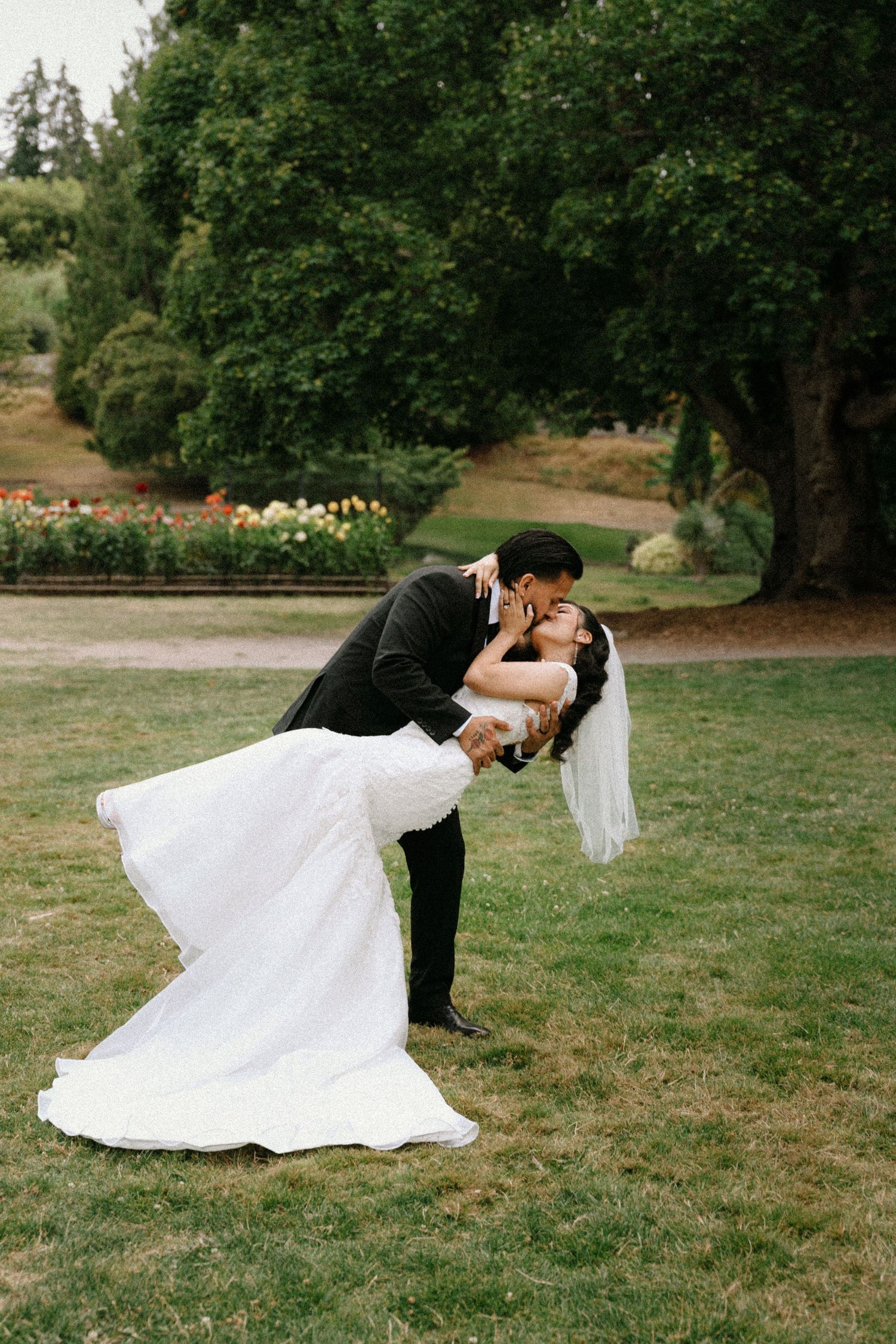 Groom dips bride for a romantic kiss in a lush green garden, bride wearing a white gown and veil.