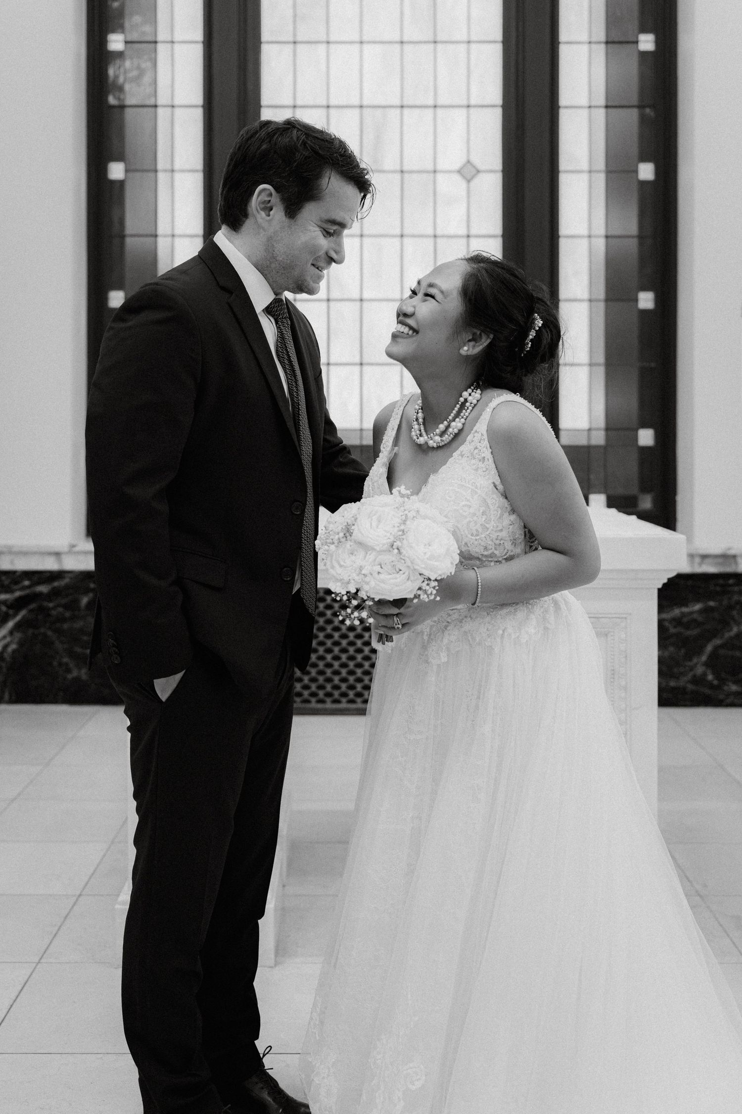 Black and white photo of a smiling bride in white gown and groom in dark suit sharing a tender moment indoors.