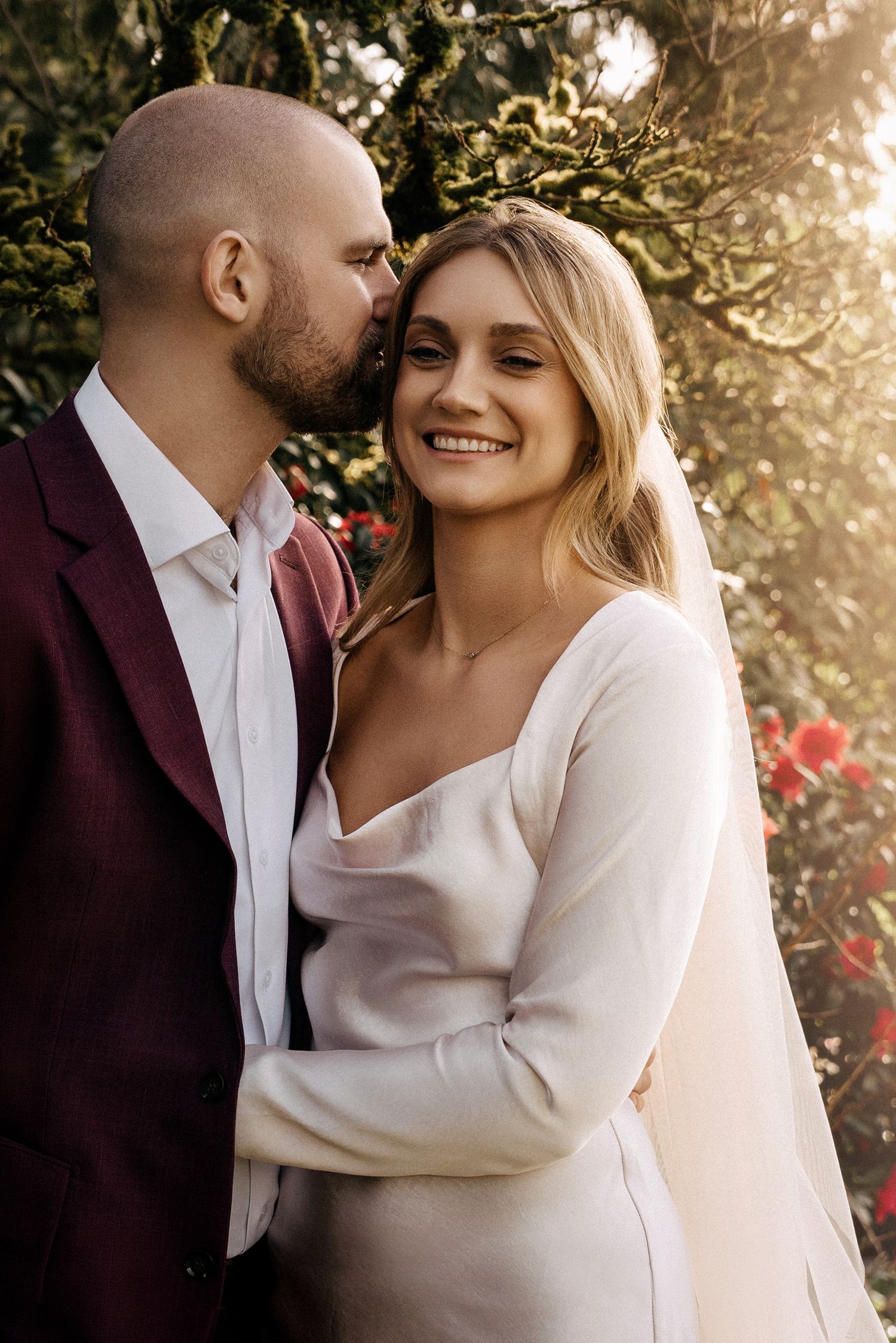 Groom in burgundy suit kissing bride on cheek as she smiles, standing outdoors in golden sunlight near blooming trees.