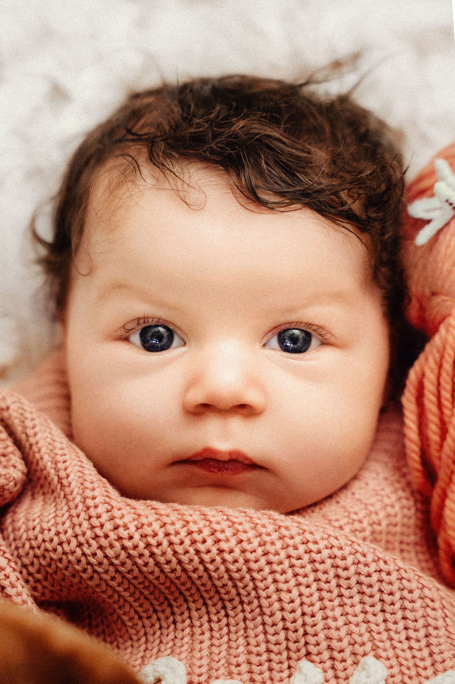Close-up portrait of a newborn baby with dark curly hair and blue eyes wrapped in a soft pink knit blanket.