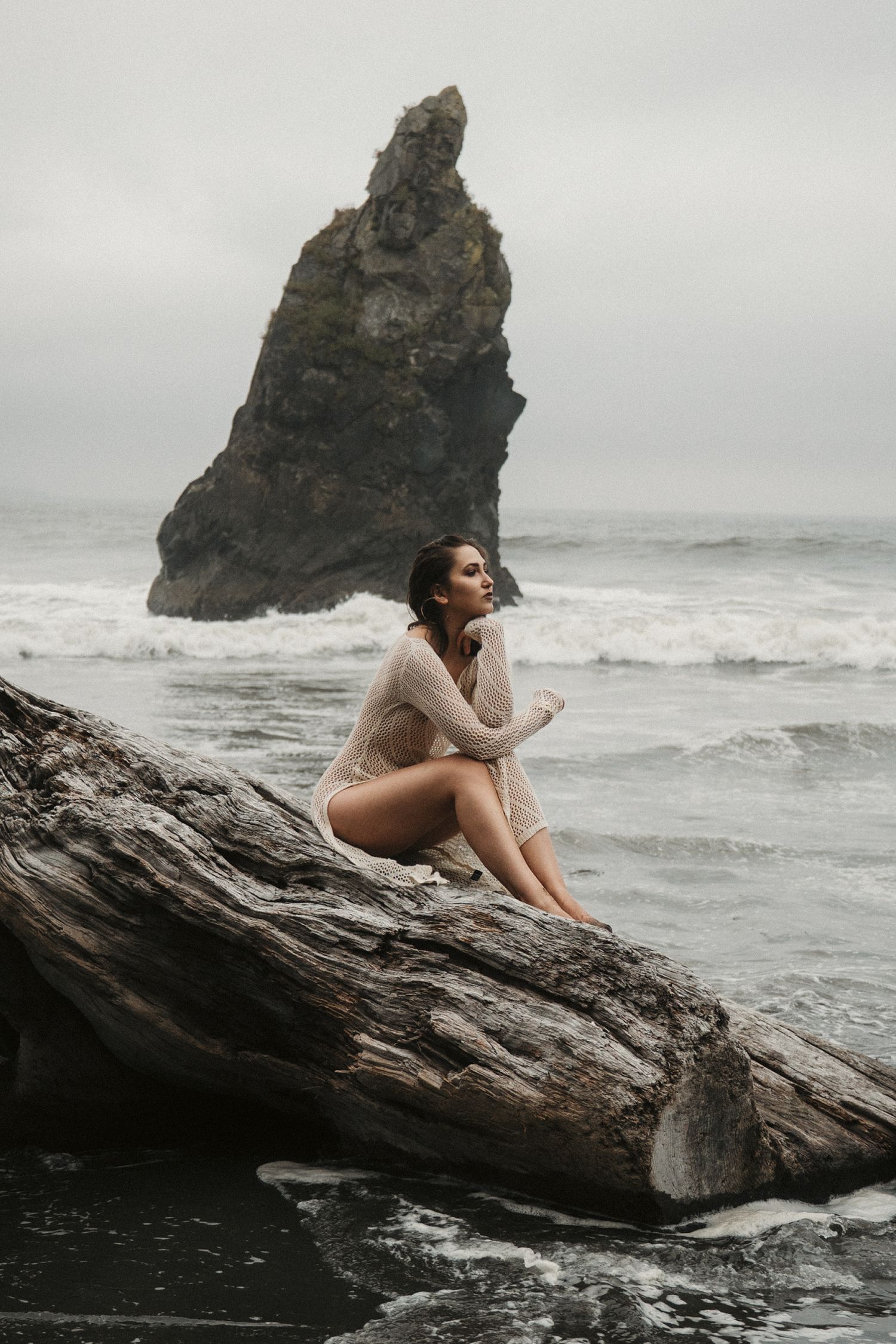 Woman in sheer cover-up sits on driftwood at moody beach with dramatic sea stack rock formation behind her.