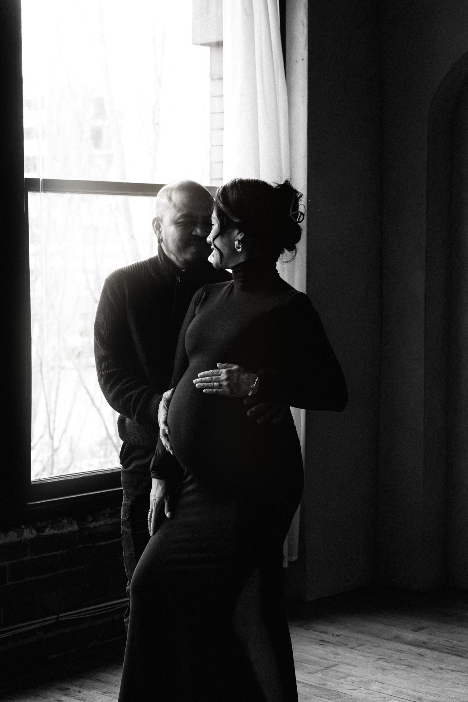 Black and white photo of a couple embracing by a window, pregnant woman in a black dress with partner behind her.