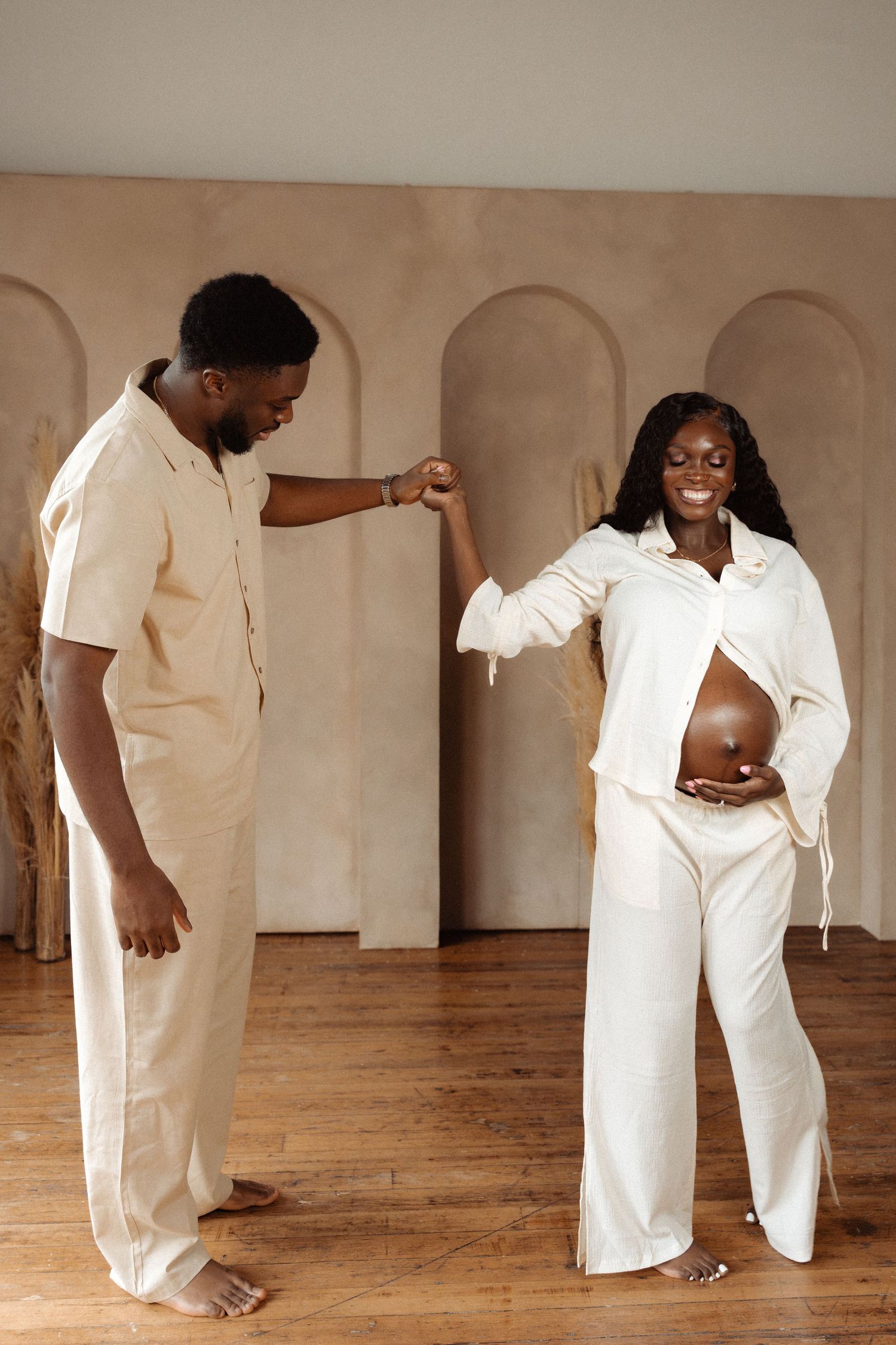 A pregnant woman in white outfit holds hands with a man in beige, both smiling in a stylish neutral-toned room.