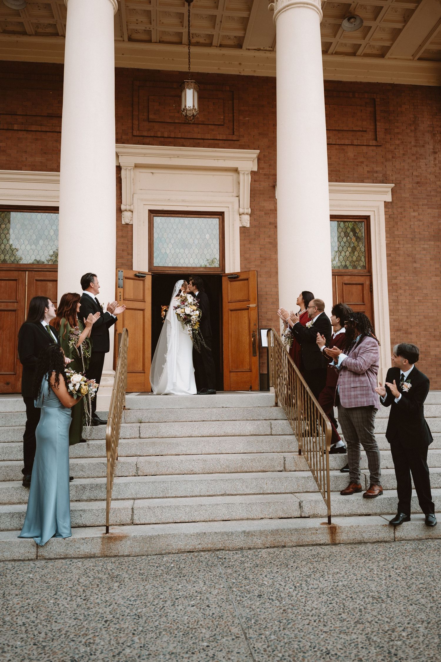 Bride and groom kiss on church steps surrounded by applauding wedding party in elegant attire.