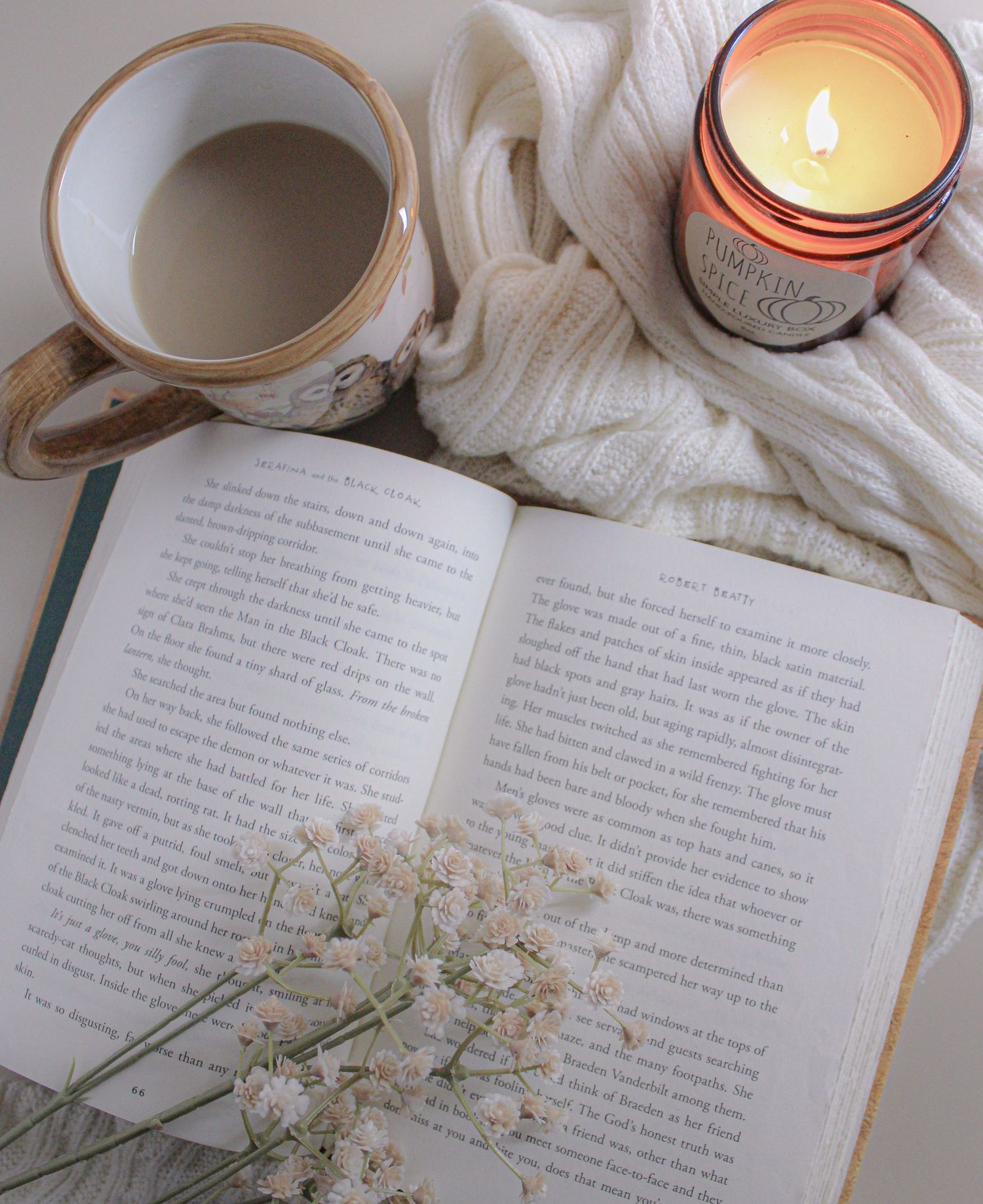 Open book with baby's breath flowers, a cozy knit blanket, lit amber candle, and a warm cup of coffee on a white surface.