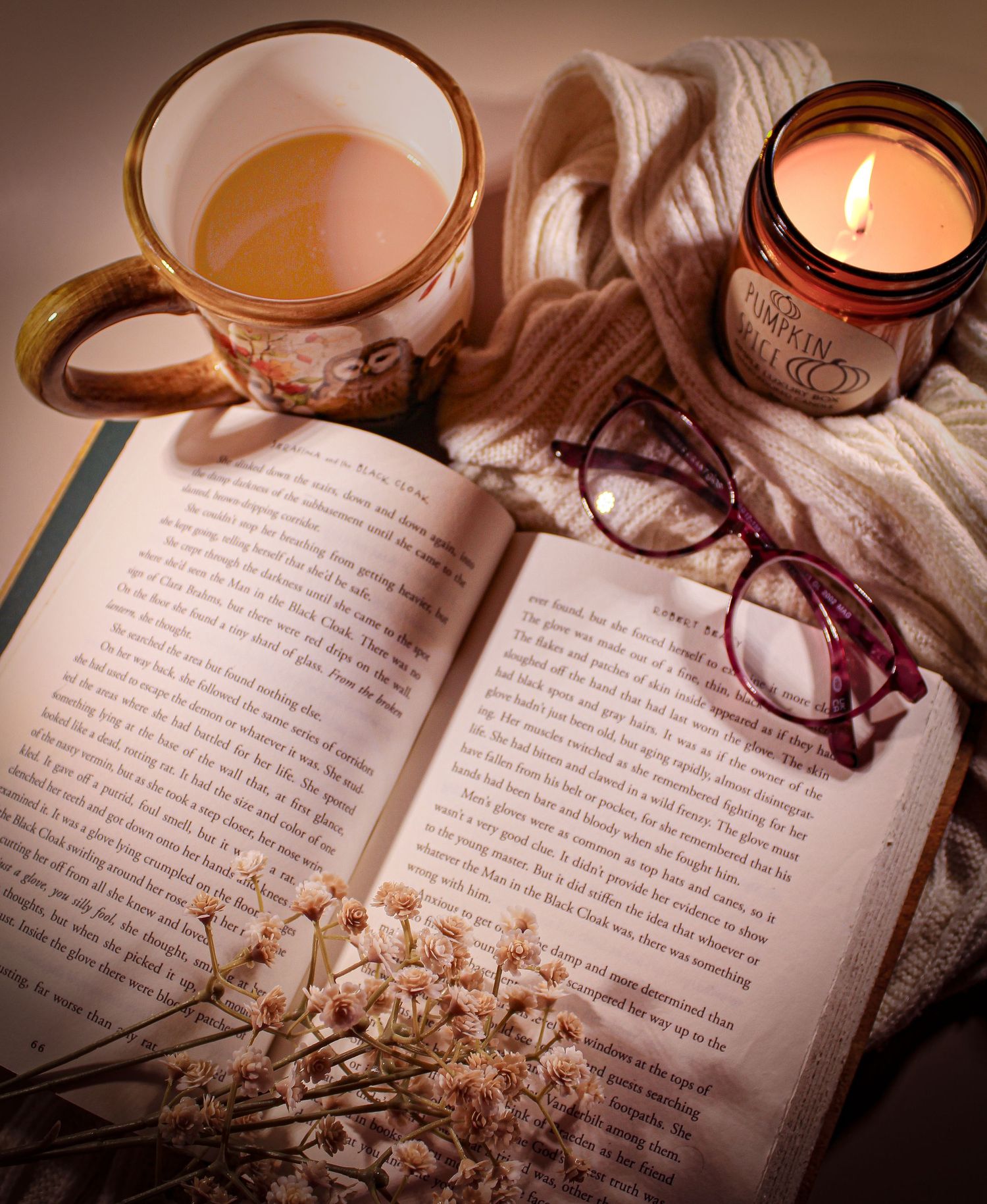 Cozy reading setup with open book, red glasses, lit candle, floral mug of tea, and dried flowers on a soft knit blanket.