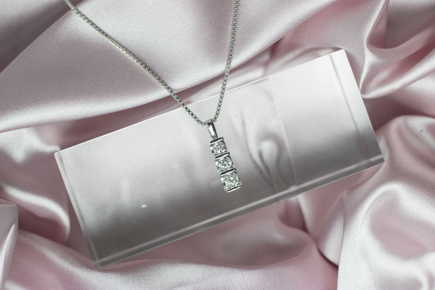 Three-stone diamond pendant necklace displayed on silver jewelry box with pink satin fabric.