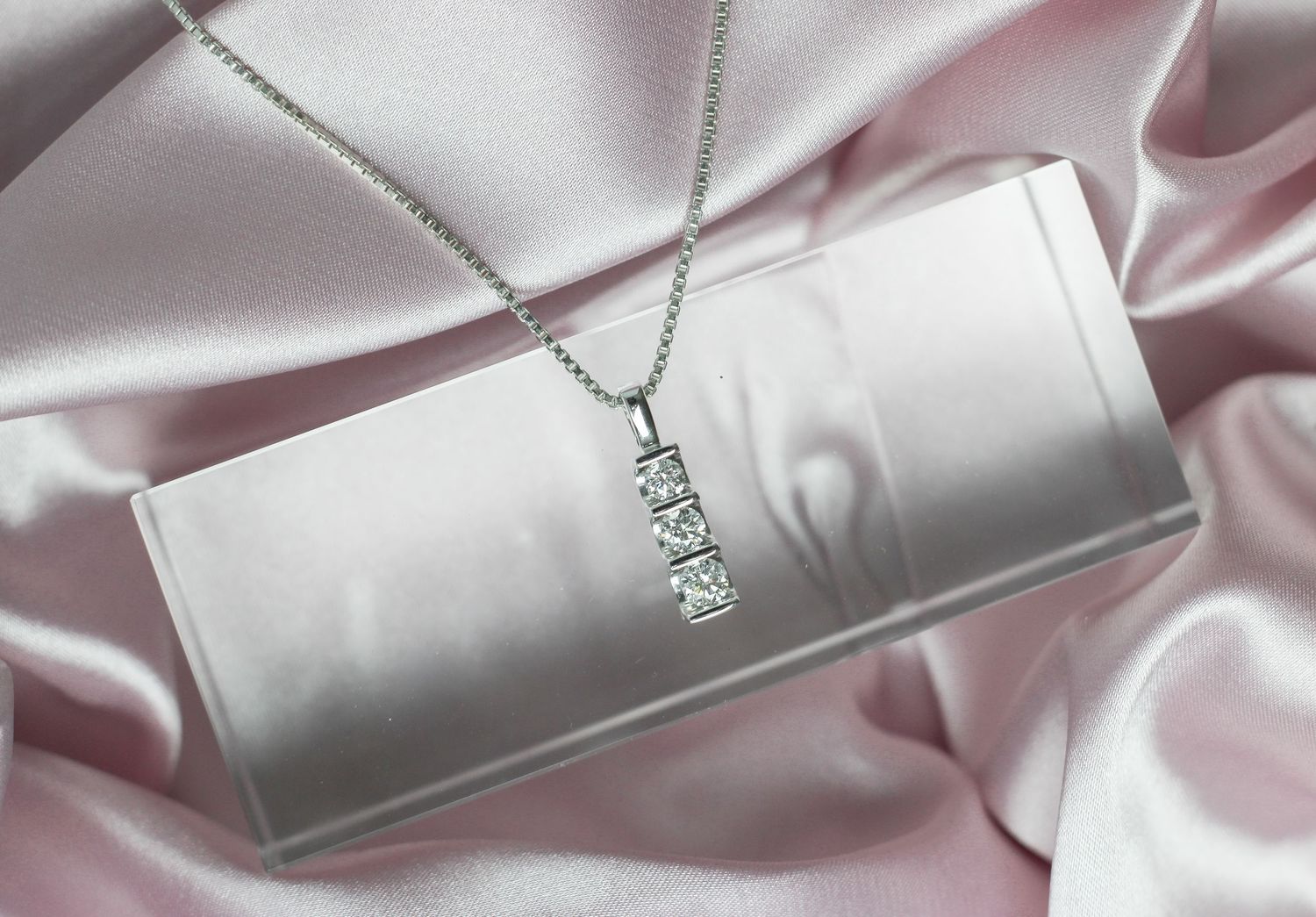 Jewelry photography. Classic three-diamond pendant necklace on silver chain atop jewelry box with pink satin fabric.