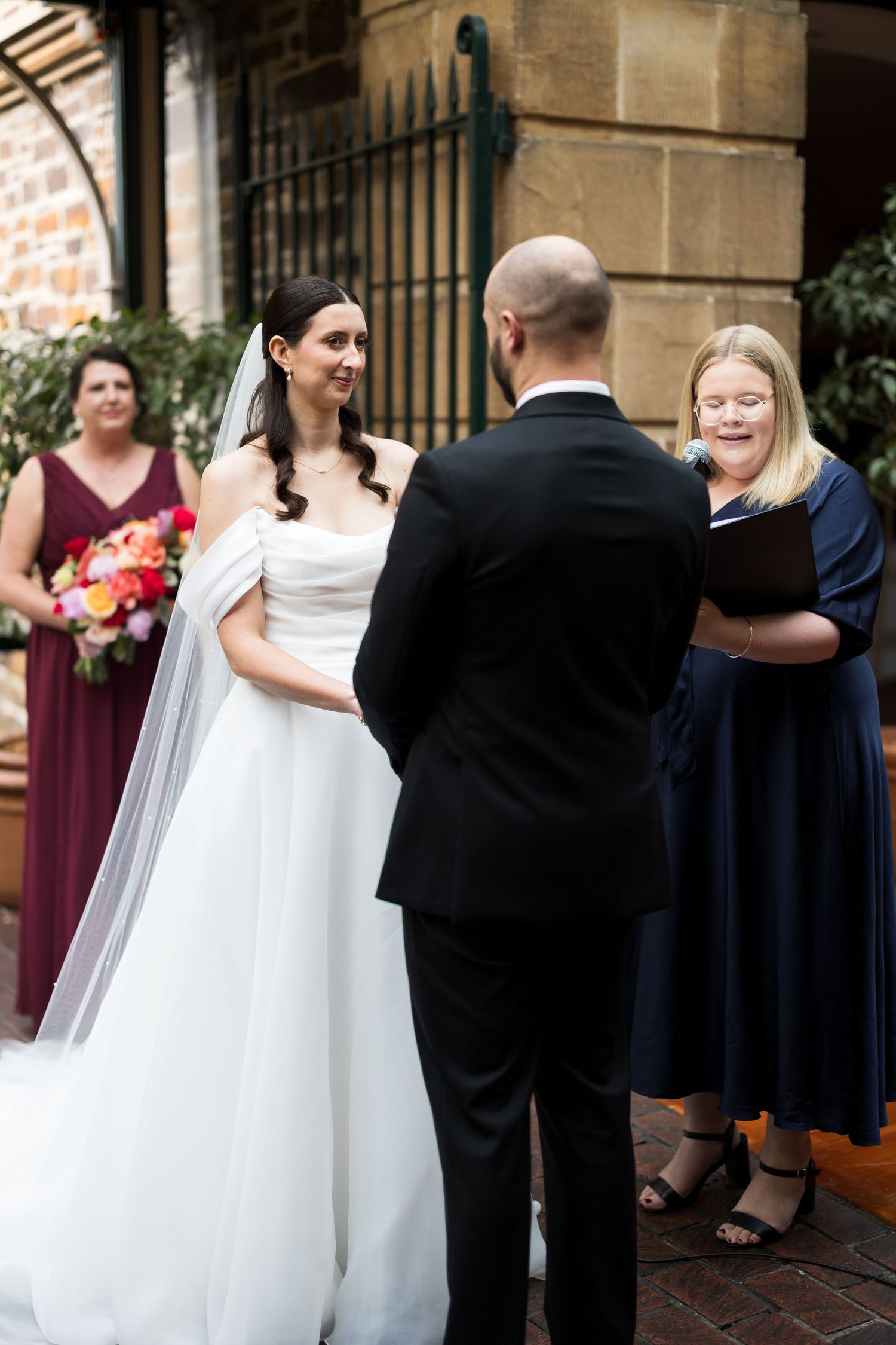Bride in white gown smiling at groom during outdoor wedding ceremony with officiant in navy dress.