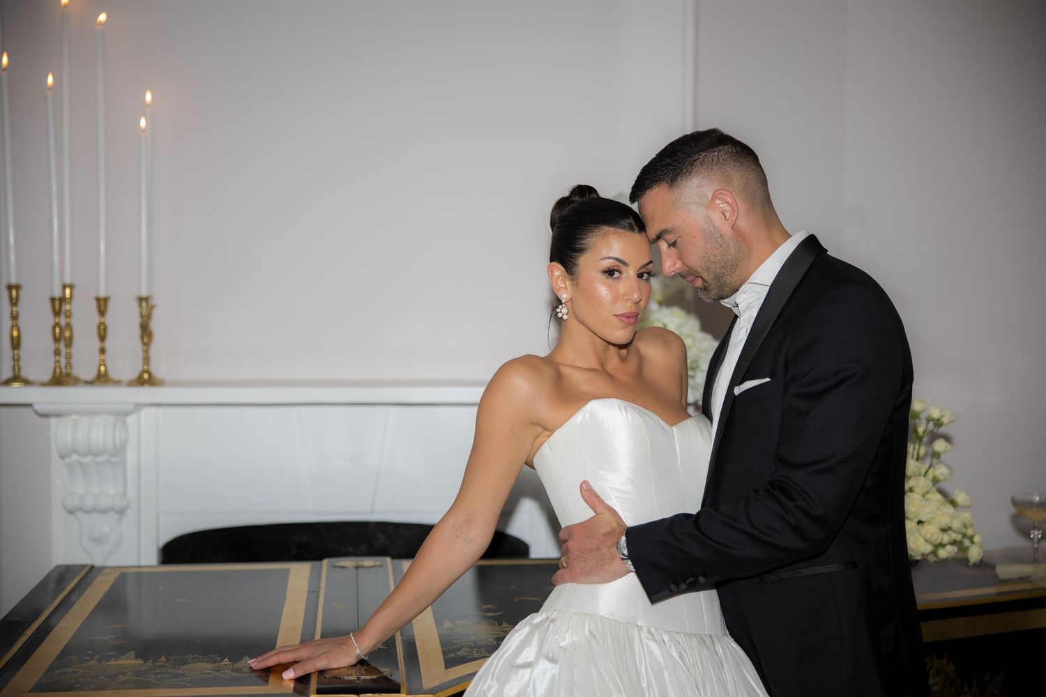 Elegant bride and groom embrace near candlelit piano in white wedding venue.
