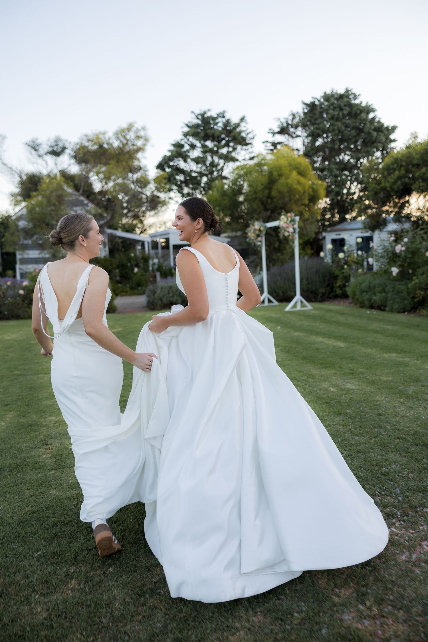 Two brides in white gowns walking hand in hand across green lawn at wedding venue.