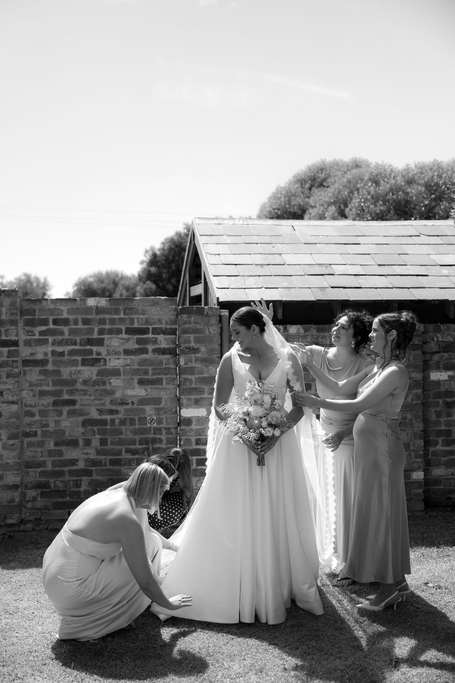 Black and white photo of bridesmaids helping bride adjust wedding dress and bouquet outdoors.