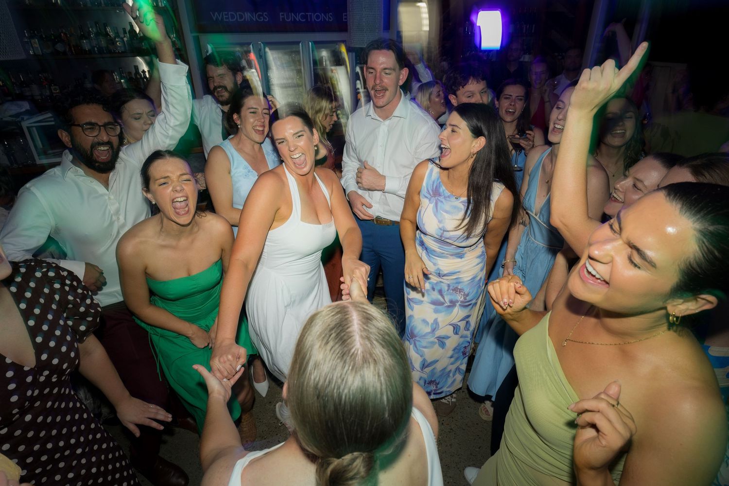 Bride in white dress dances joyfully with guests on a crowded wedding reception dance floor.