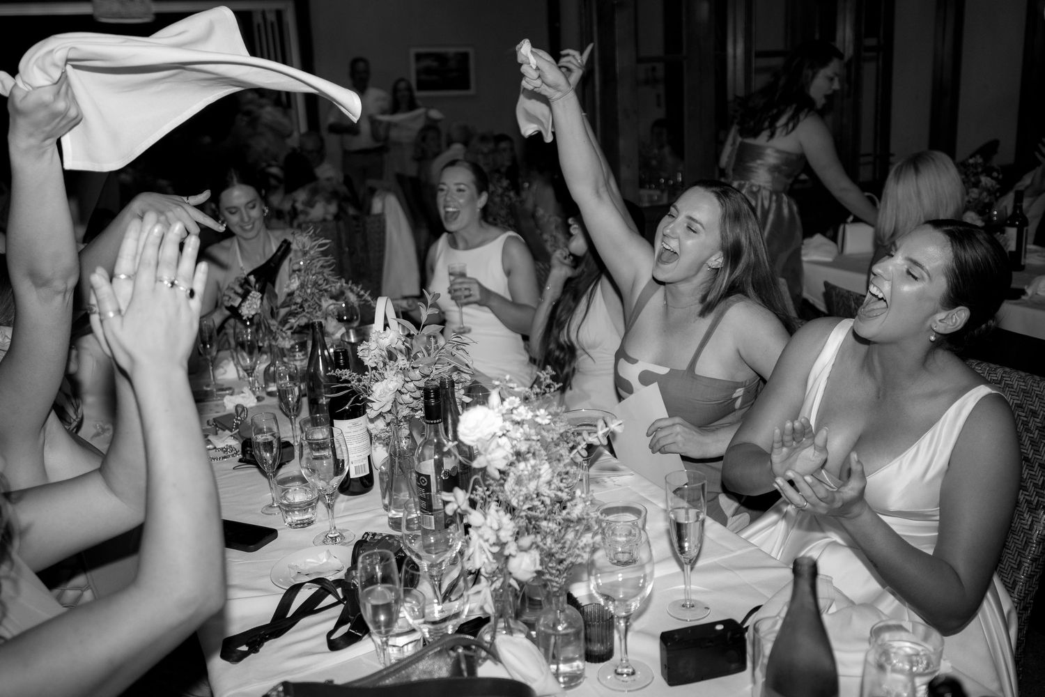 Black and white photo of joyful wedding guests celebrating at a reception dinner table.