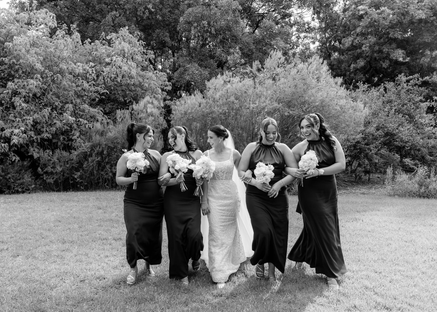 Black and white photo of bride and bridesmaids in dark gowns walking outdoors.