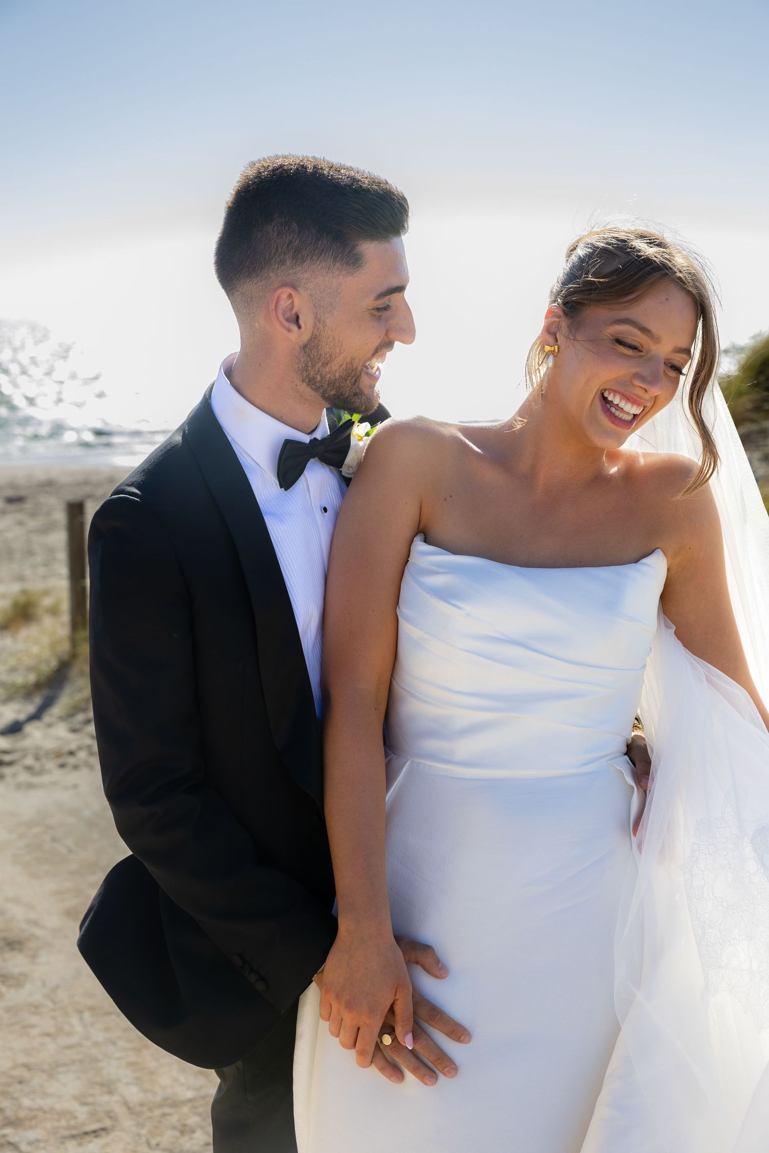 Happy bride in strapless white gown holds hands with groom in black tuxedo at sunny outdoor wedding.