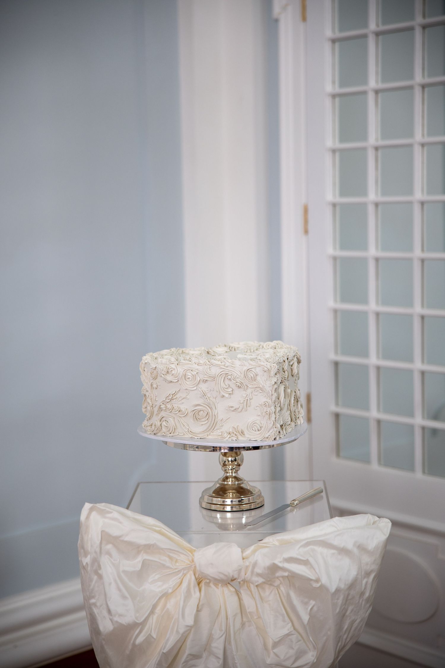 Single-tier white wedding cake with rosette frosting on a gold stand, displayed near a French door with a bow-tied chair.
