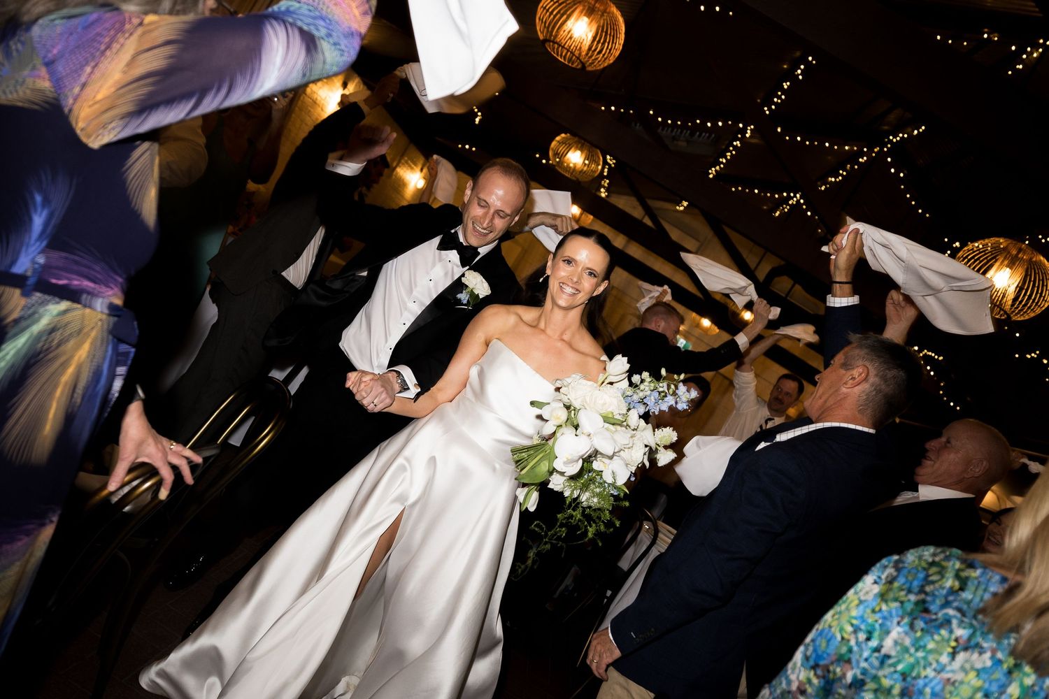 Smiling bride in a white gown holding a bouquet walks hand in hand with her groom as wedding guests wave white napkins around them.