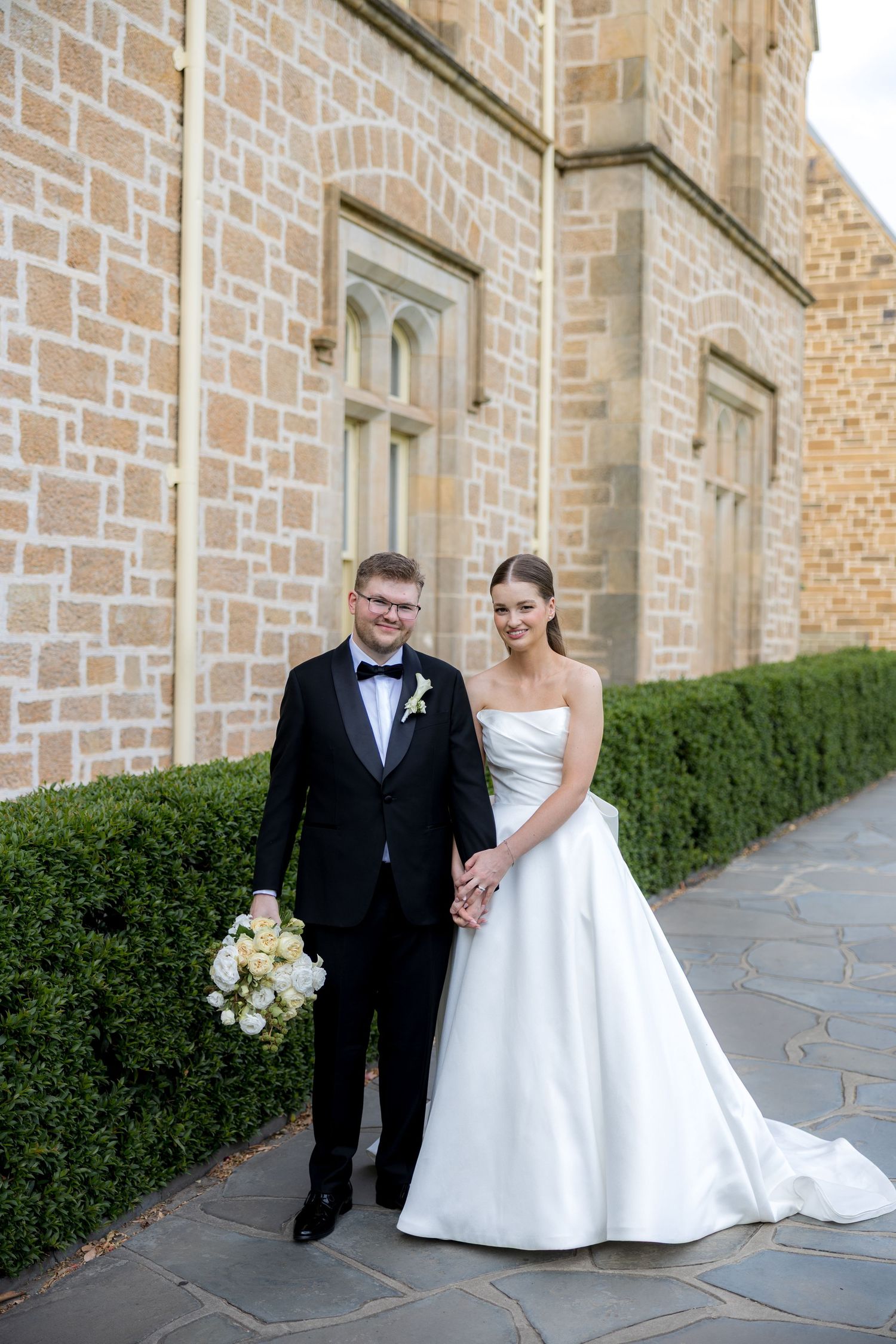 Bride in white strapless gown and groom in black tuxedo hold hands smiling outside a stone building.