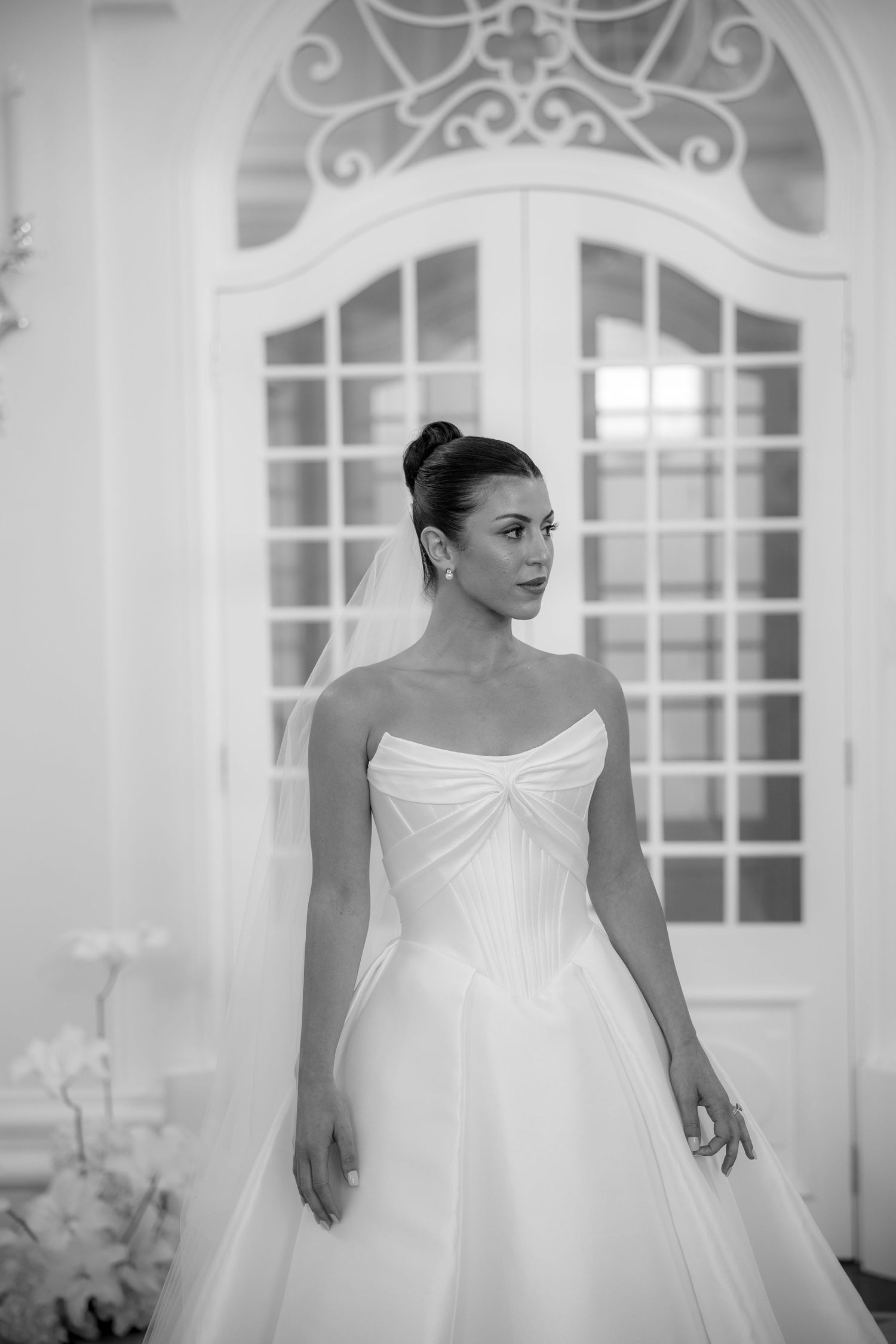 Black and white portrait of a bride in a strapless ballgown with veil standing before ornate French doors.