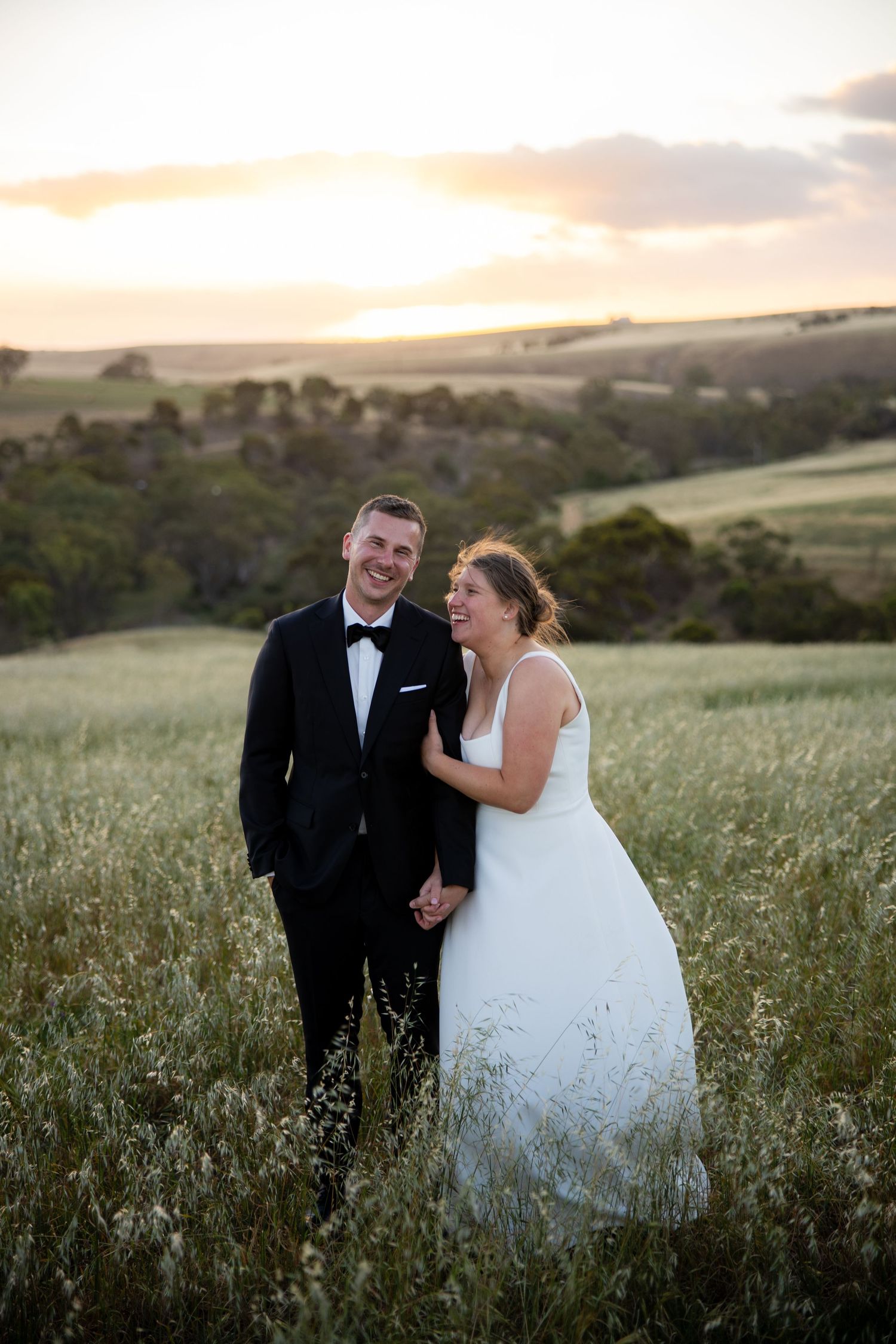 Laughing bride and groom stand in golden field at sunset during wedding portraits.