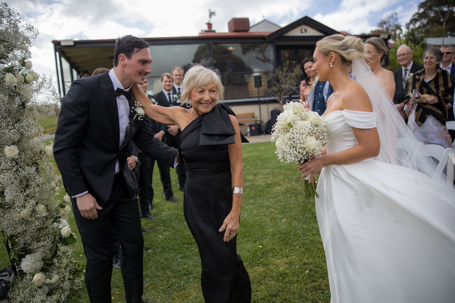Older woman in black dress congratulates bride and groom during outdoor wedding ceremony at vineyard.