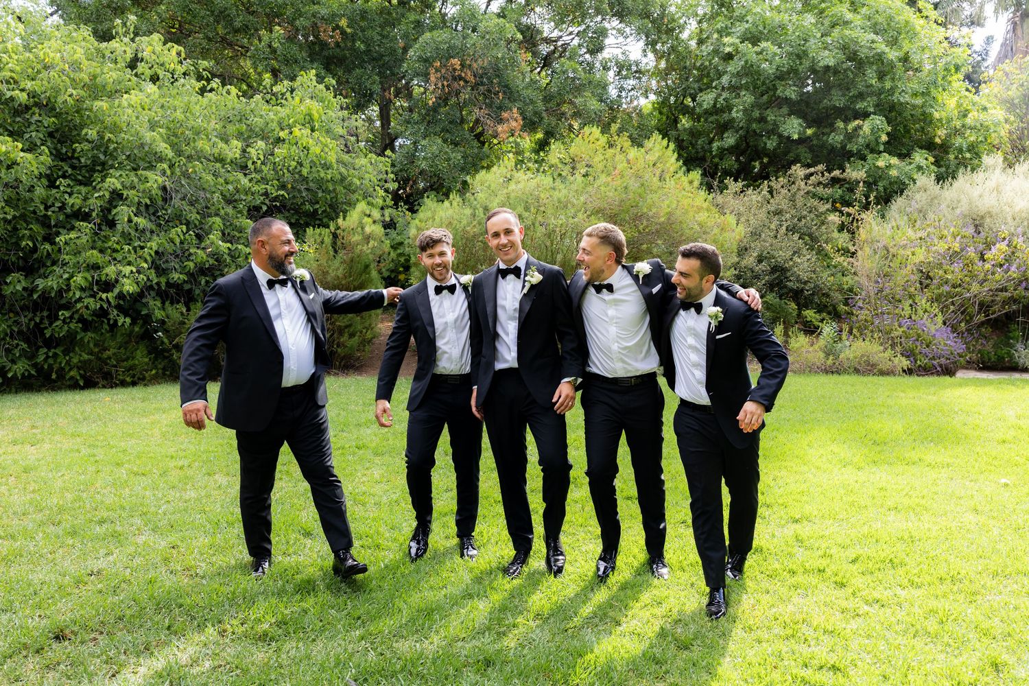 Five groomsmen in black tuxedos laughing together on green lawn with lush garden backdrop.