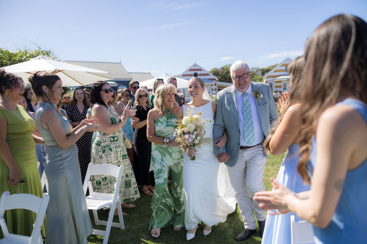 Bride walking down outdoor aisle with parents while guests applaud at sunny garden wedding.