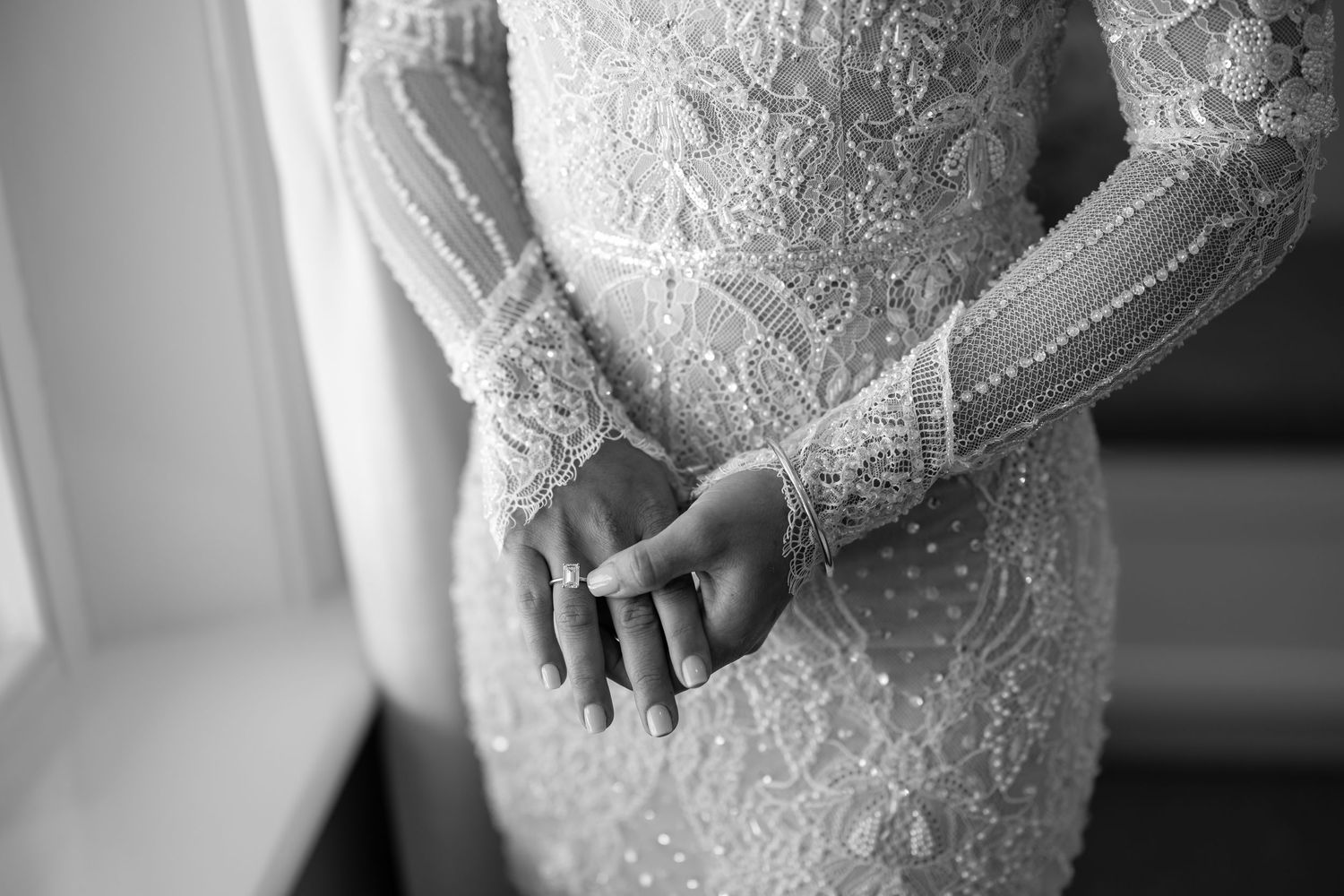 Black and white photo of a bride in a lace wedding gown with pearl buttons, hands clasped at her waist.