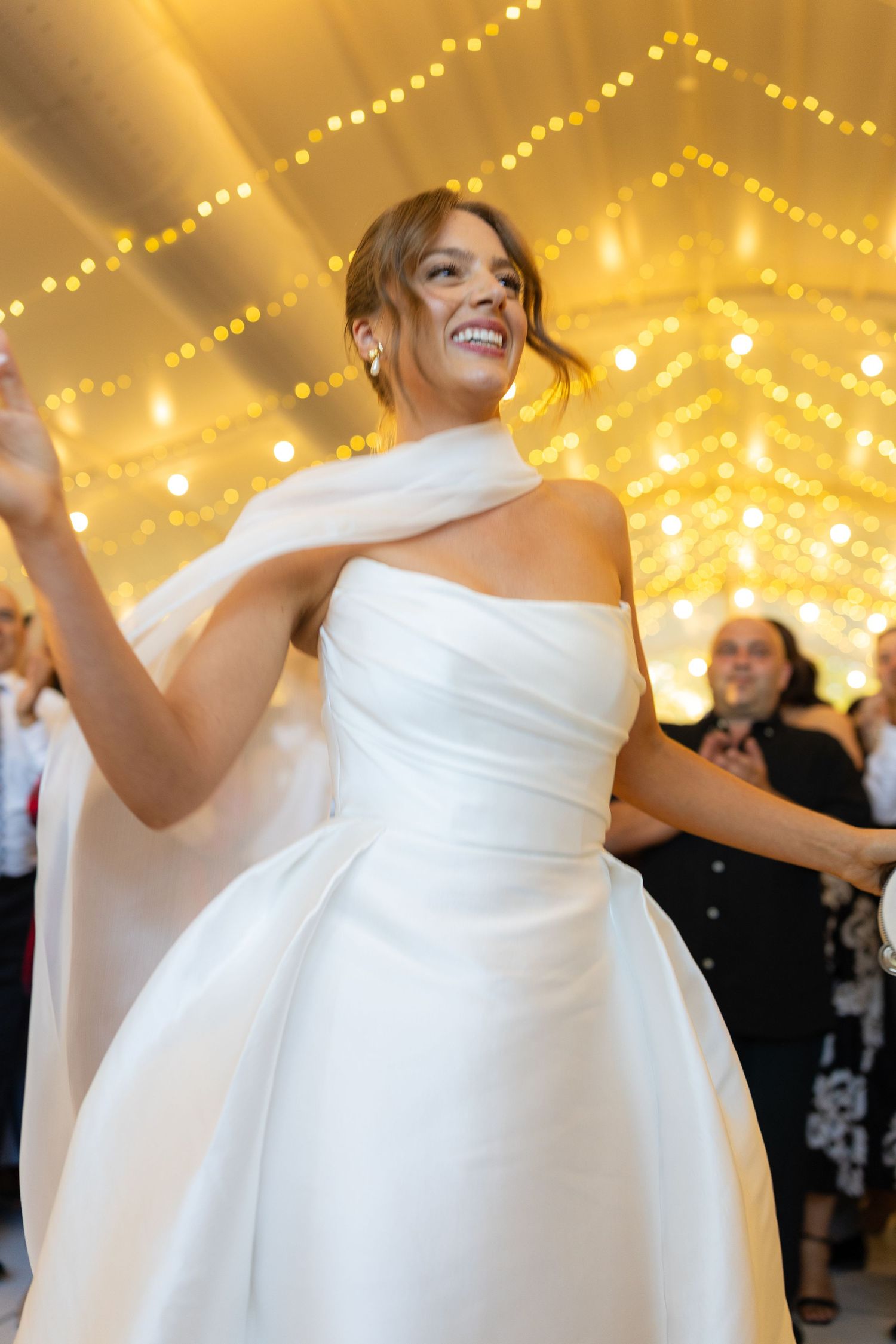 Radiant bride in strapless white ball gown with scarf dances under warm fairy lights at reception.