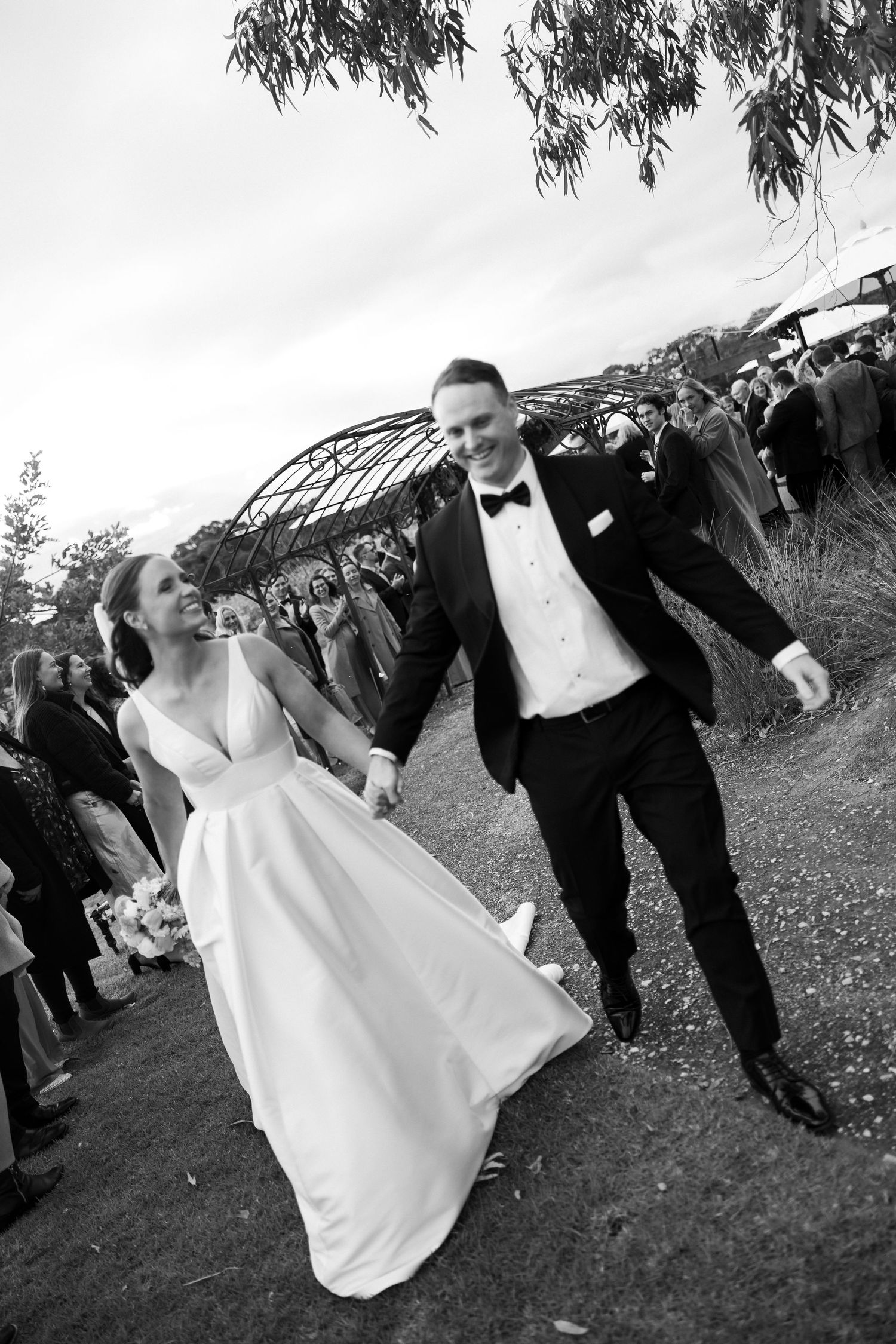 Black and white photo of a joyful bride and groom walking hand in hand past wedding guests outdoors.