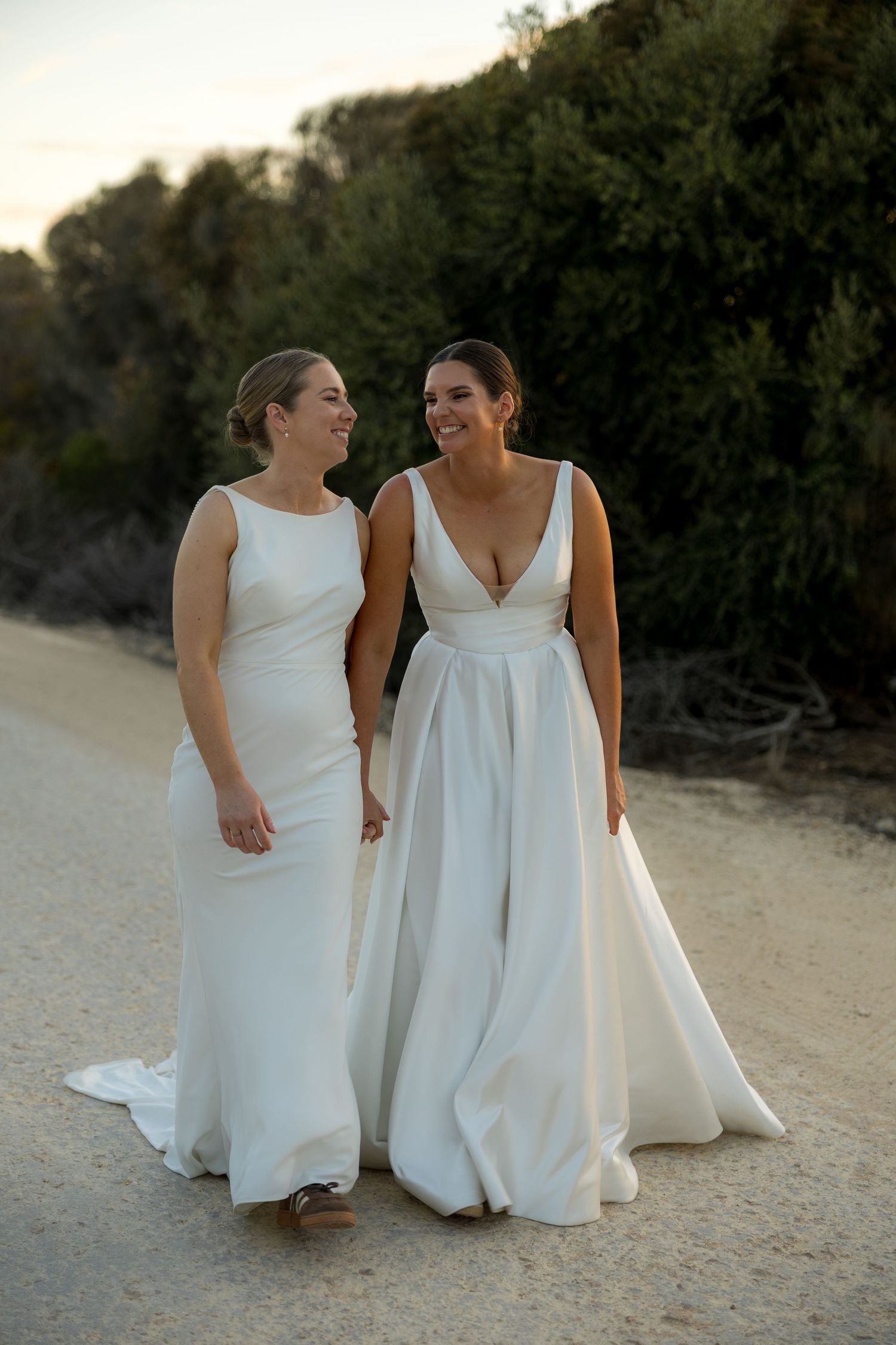Two brides in elegant white wedding gowns walking hand in hand on a sandy path at sunset.